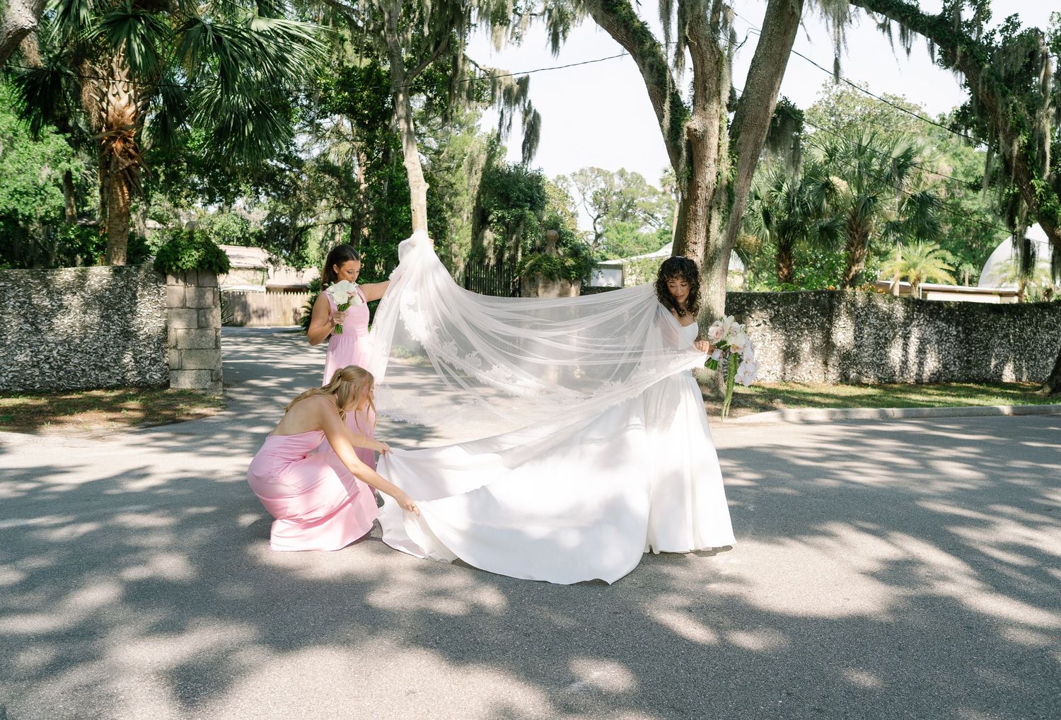 A flowing white bridal veil billows in the breeze under Spanish moss draped trees at an outdoor garden venue.