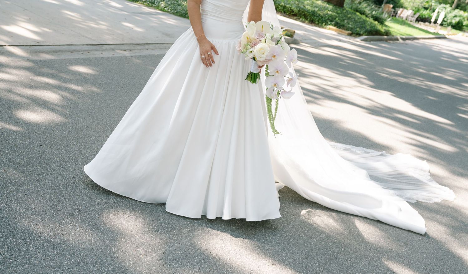 A flowing white wedding dress with a long train photographed on a sunny pathway.