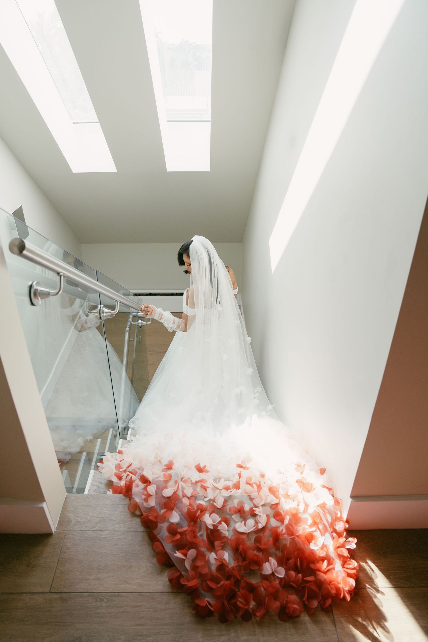Bride in white gown with coral ombre train descending modern staircase.
