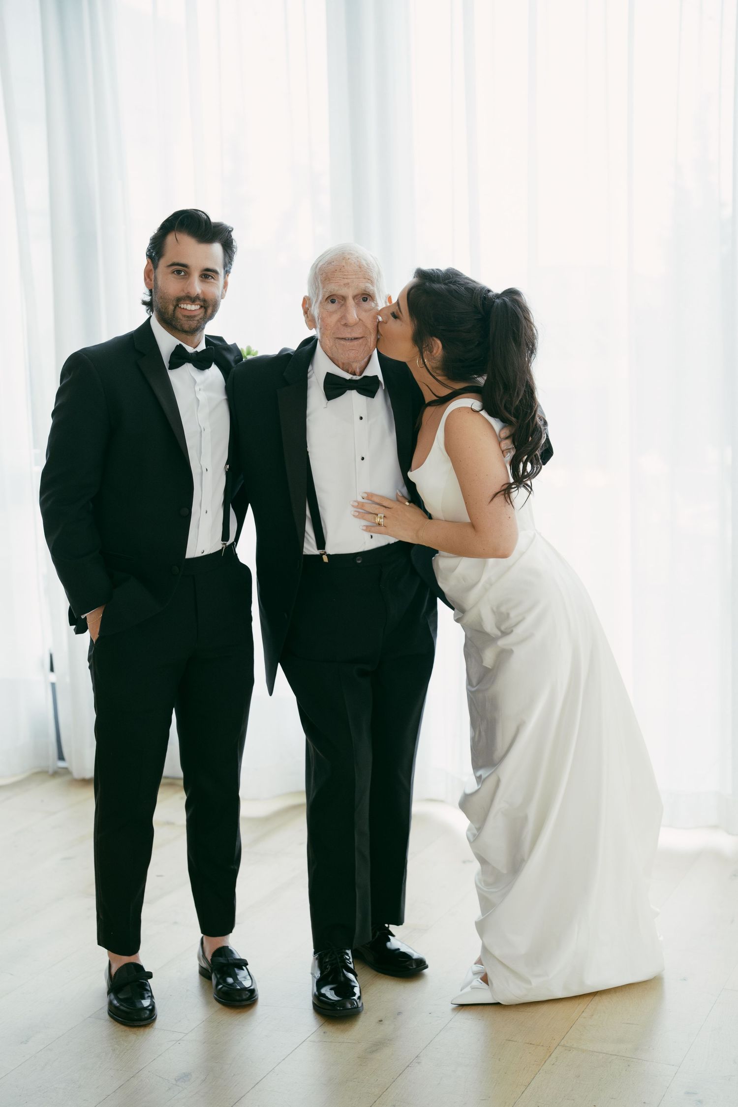 Three people in formal attire share a heartwarming moment in a bright white room.