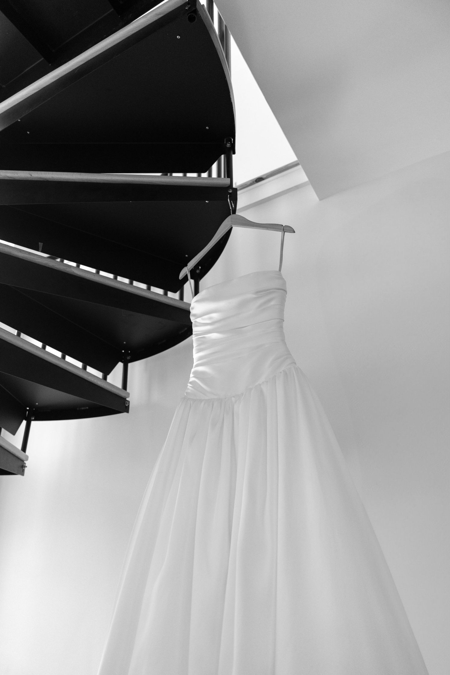 Black and white photo of an elegant wedding dress hanging on a spiral staircase.