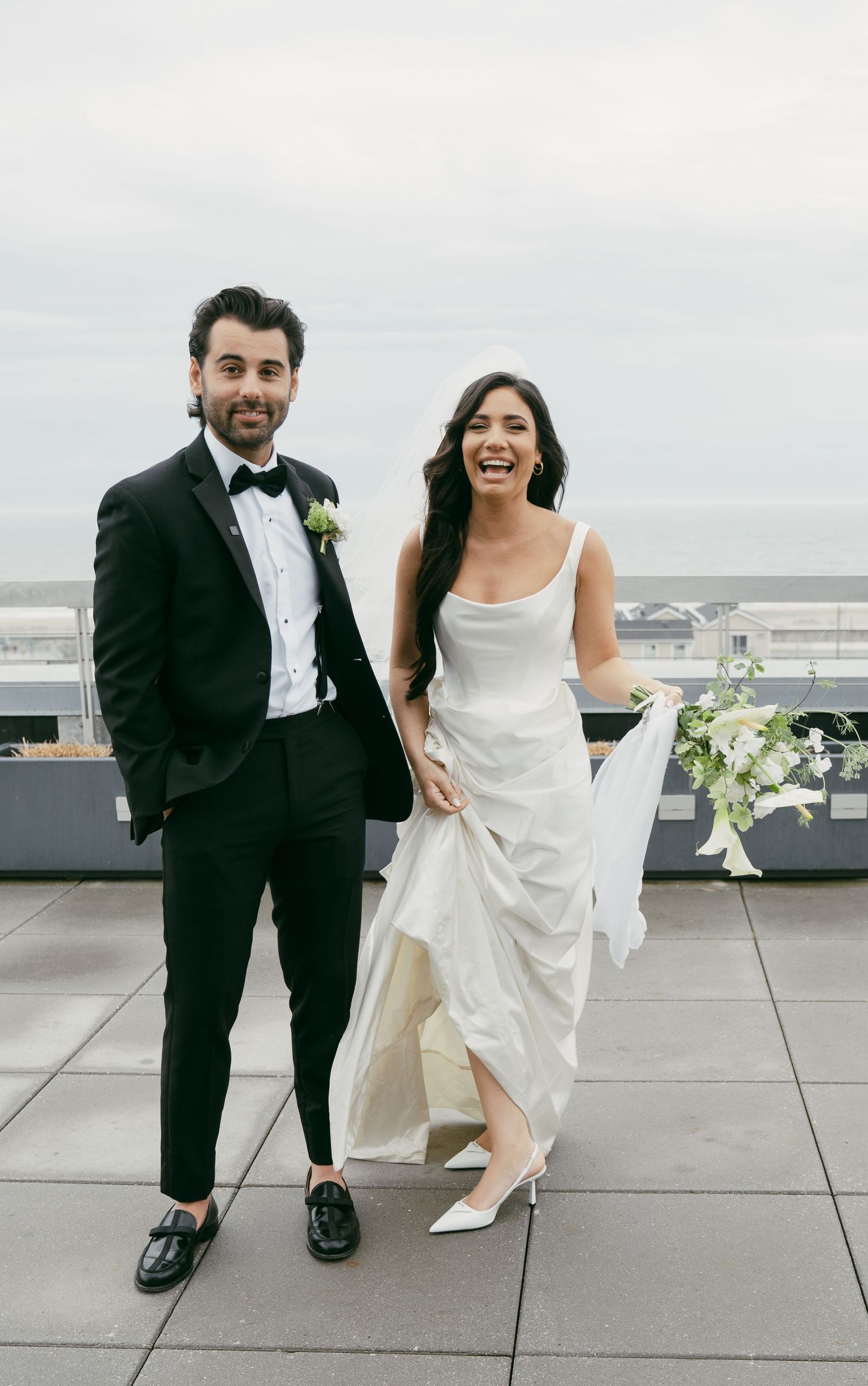 A joyful wedding couple poses outdoors on a rooftop terrace with white floral decorations and cityscape views.