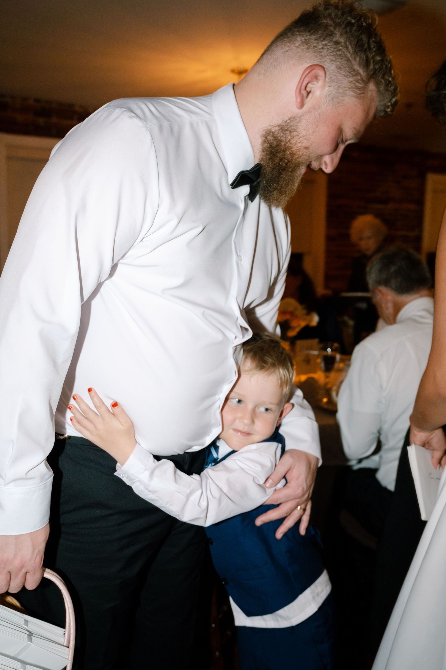 A tender moment between a bearded man in formal wear and a young child at a wedding reception.