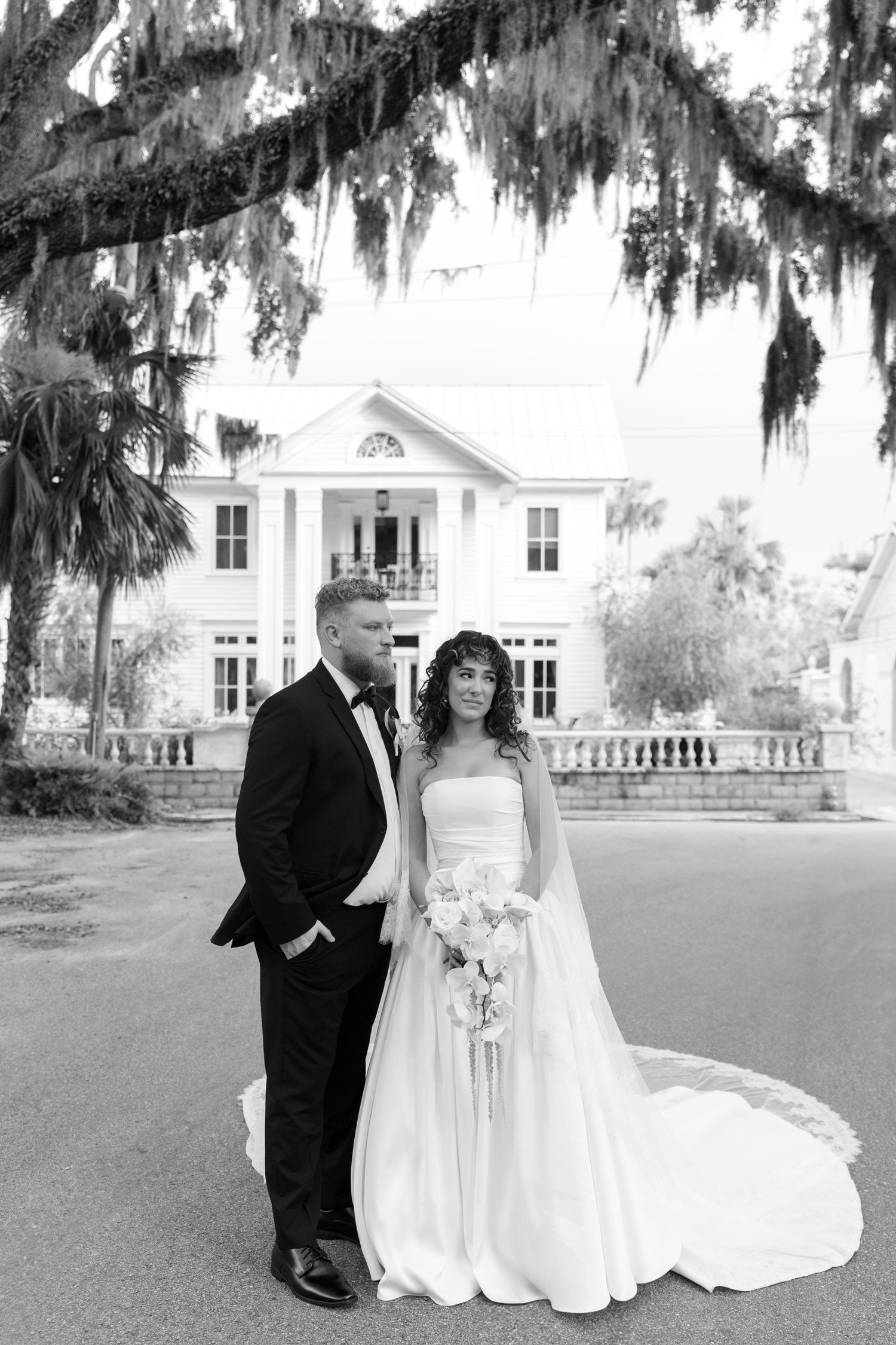 Black and white wedding portrait with couple standing in front of elegant white colonial mansion.