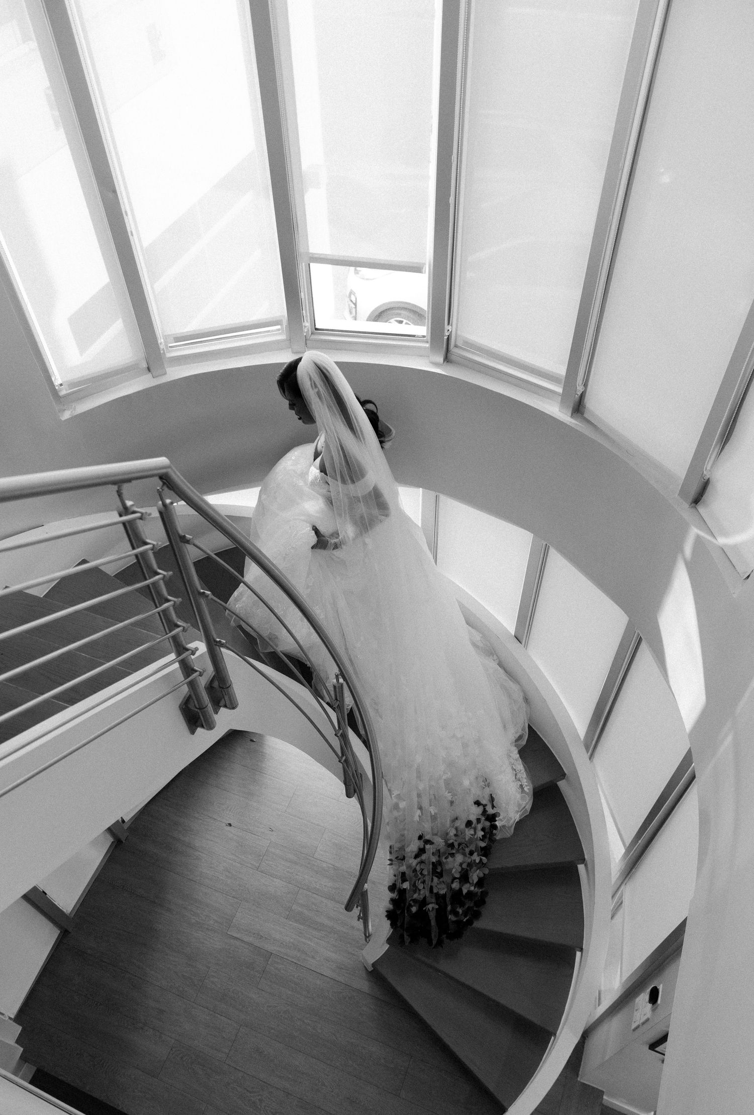 Black and white photo of bride descending spiral staircase in flowing gown.