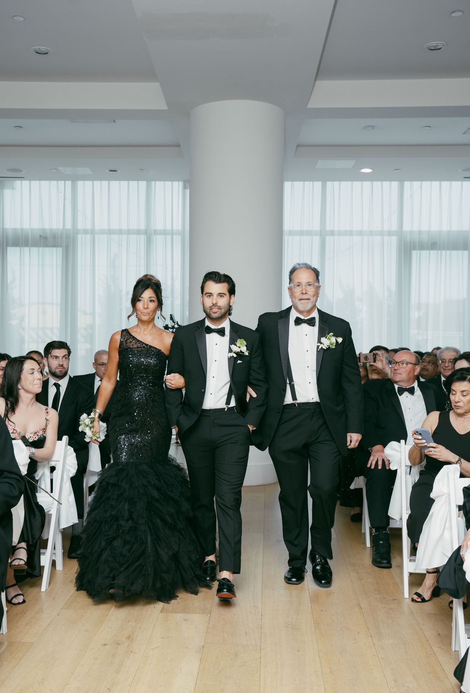 Wedding party members walk down the aisle in formal black attire during an elegant indoor ceremony.