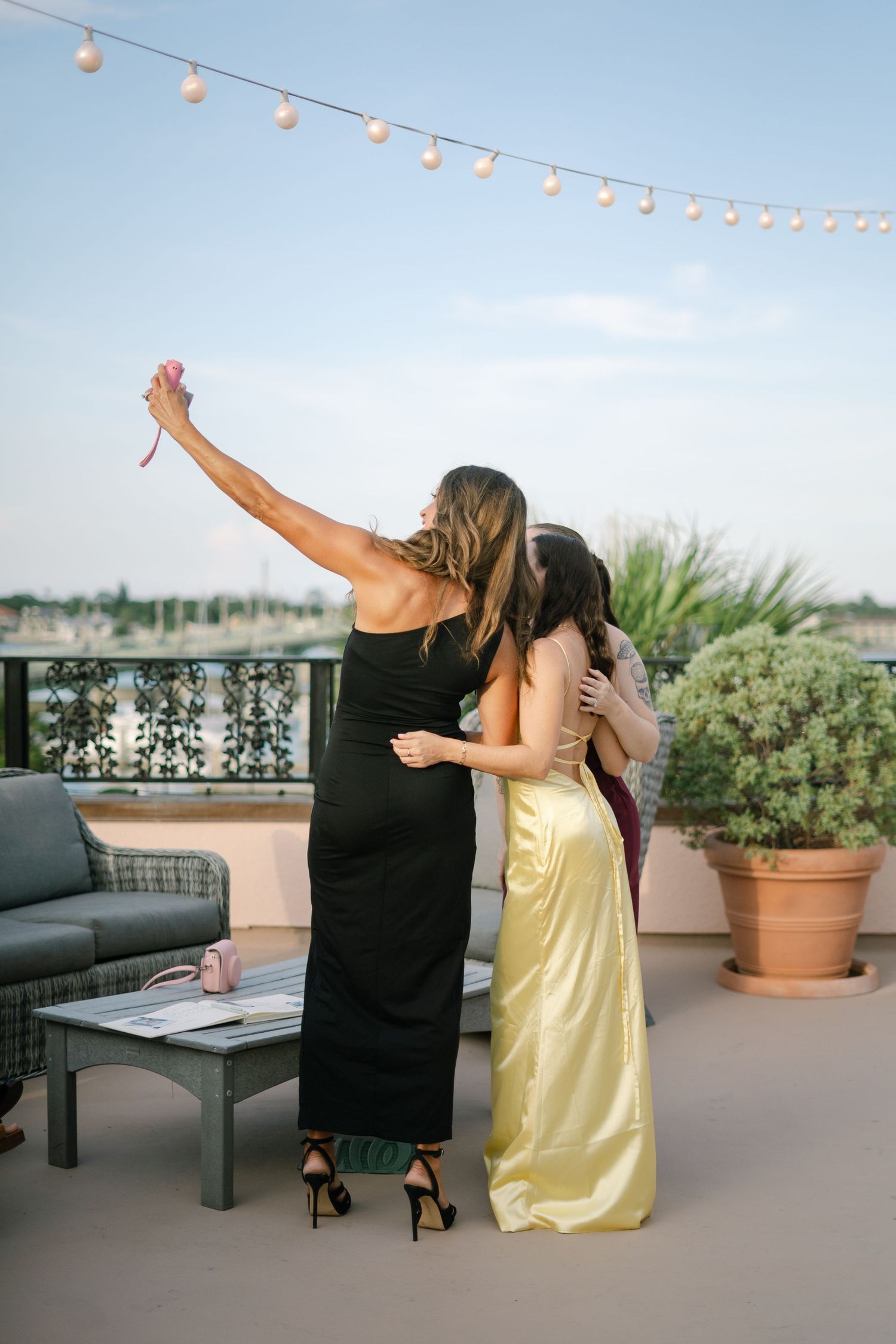 Two women in formal dresses dancing together on a rooftop patio under string lights at sunset.
