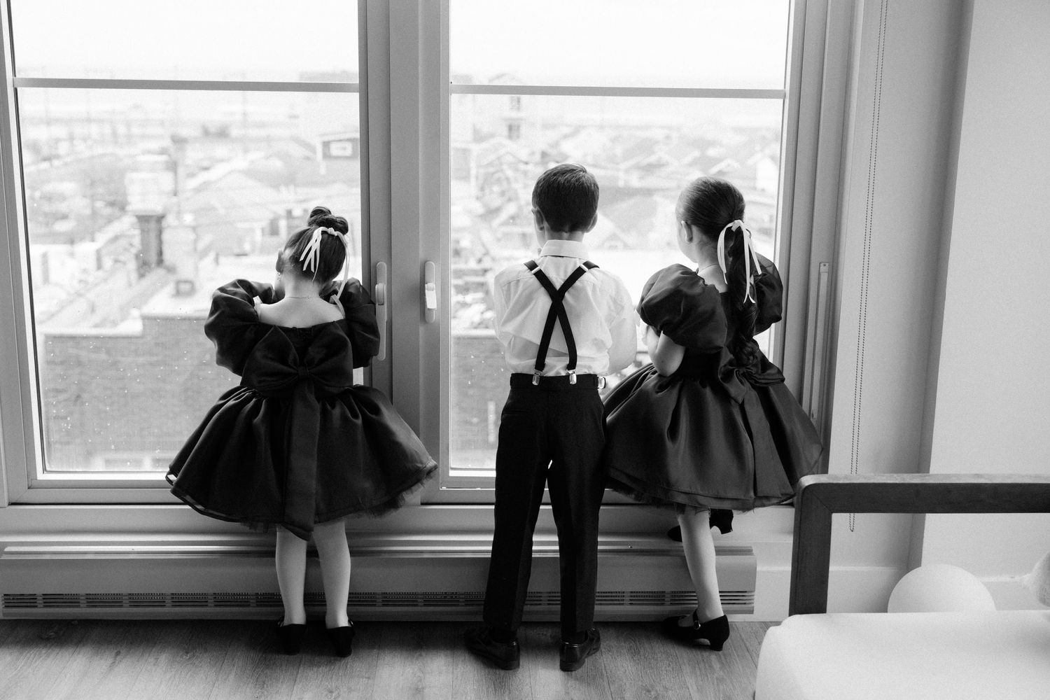 Three children in vintage clothing look out large windows at a cityscape view from high up in black and white.