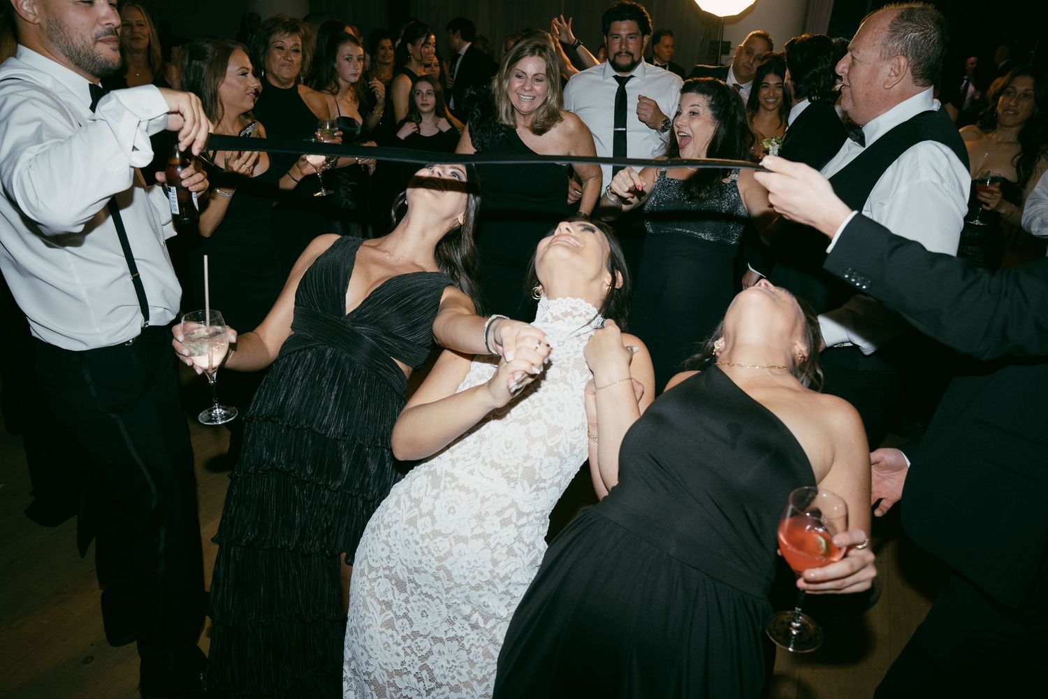 Wedding party dances and celebrates energetically on a dark dance floor at a reception.