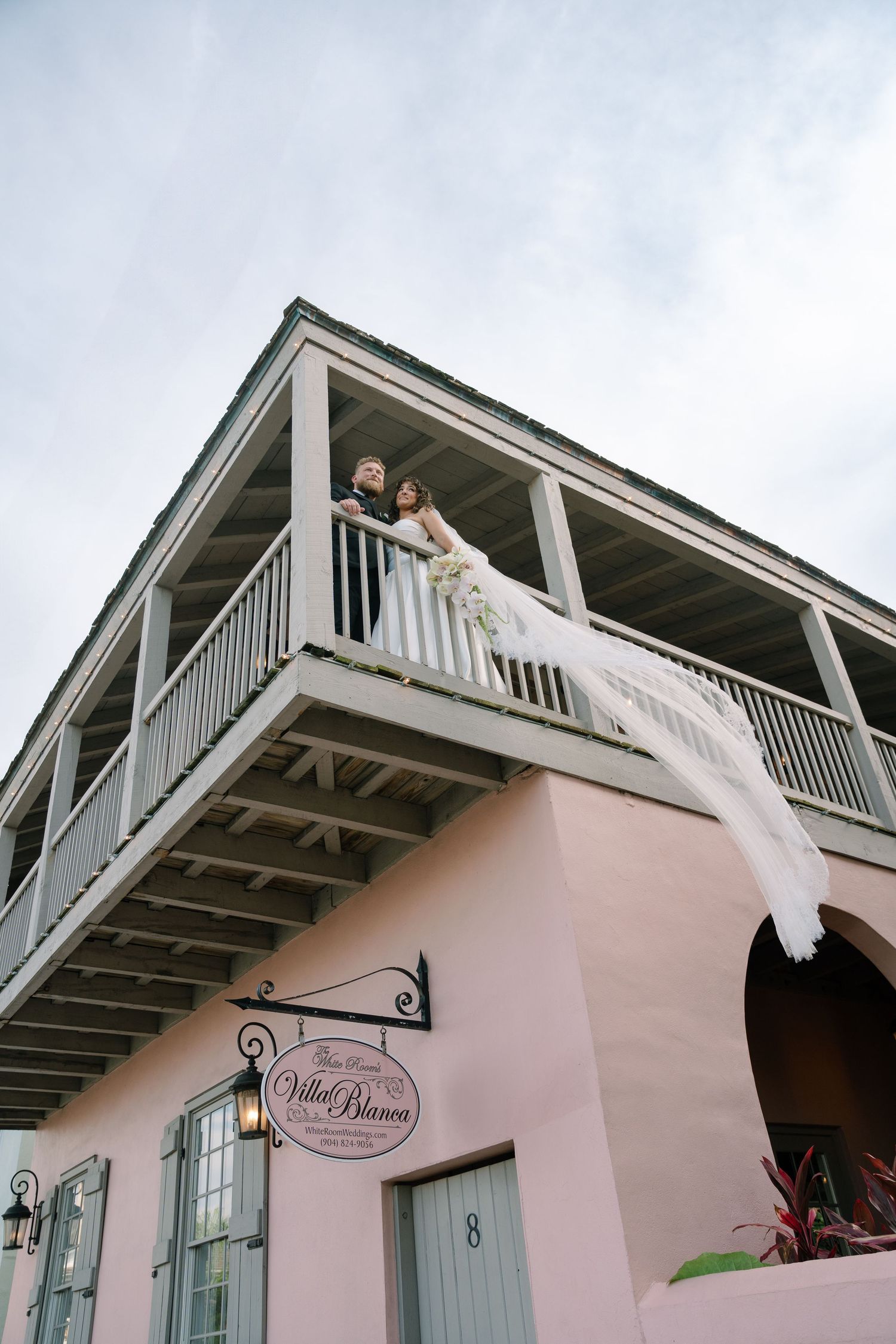 A beautiful pink villa with white trim balcony where a wedding couple stands overlooking the street below.