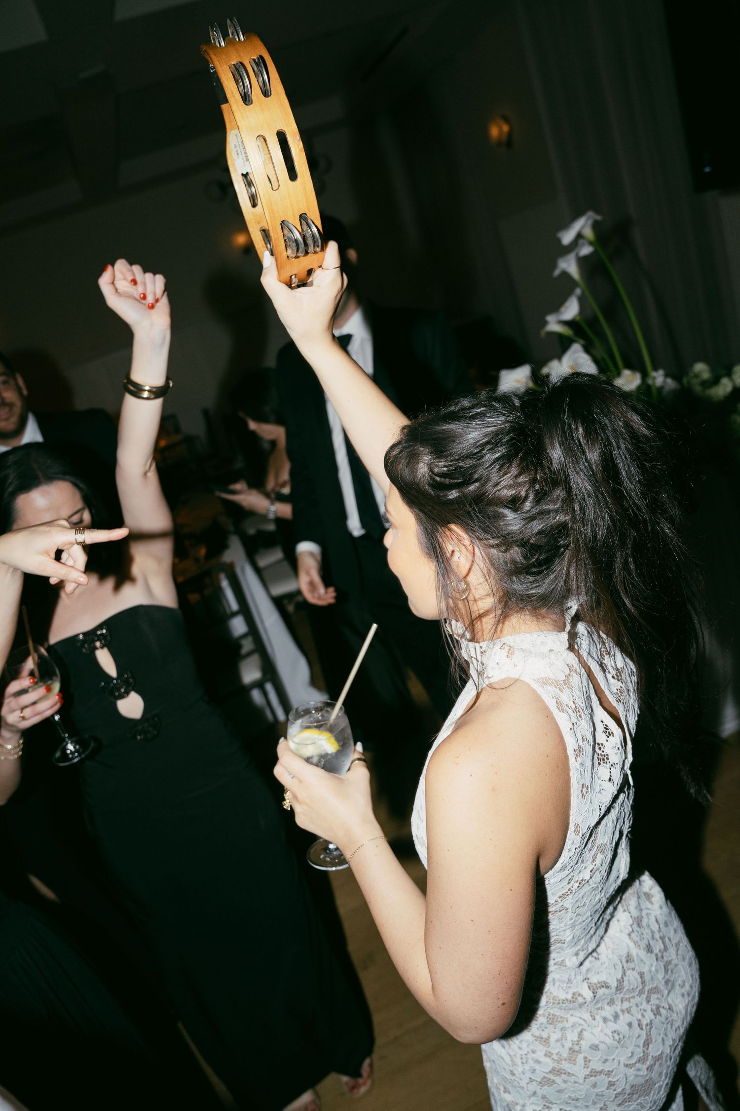 Wedding celebration on a dark dance floor with people holding up tambourines and drinks.