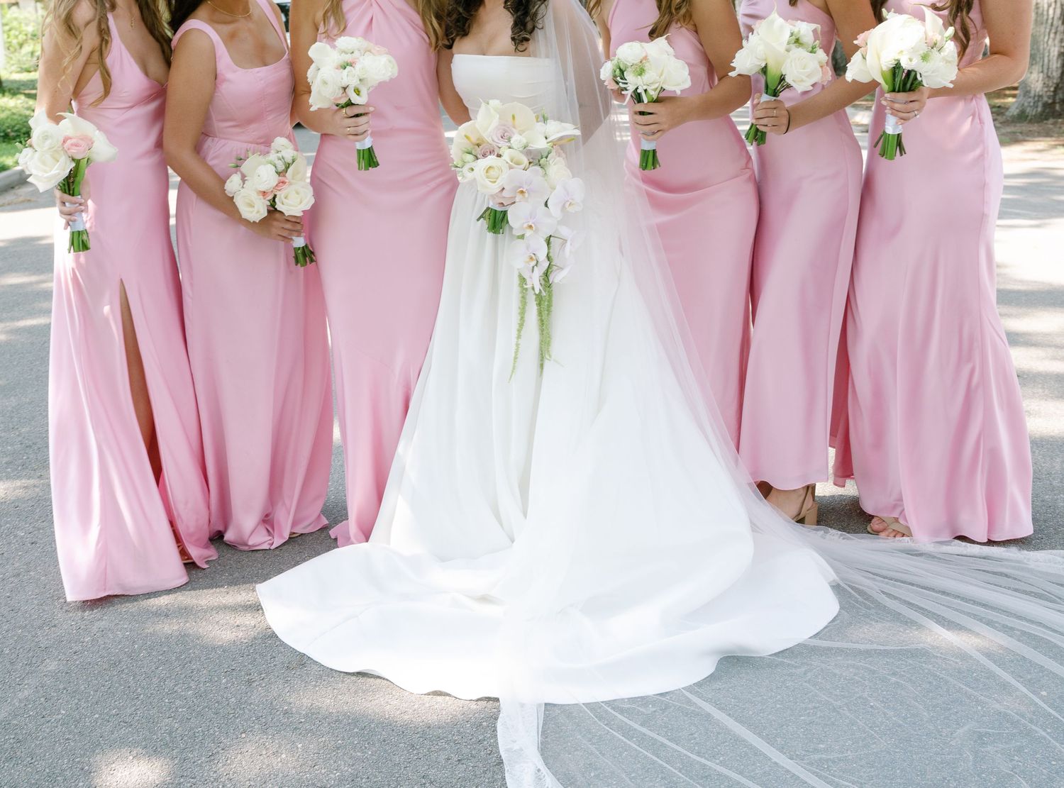 Bridesmaids in pink floor-length dresses standing with bride in white gown outdoors.