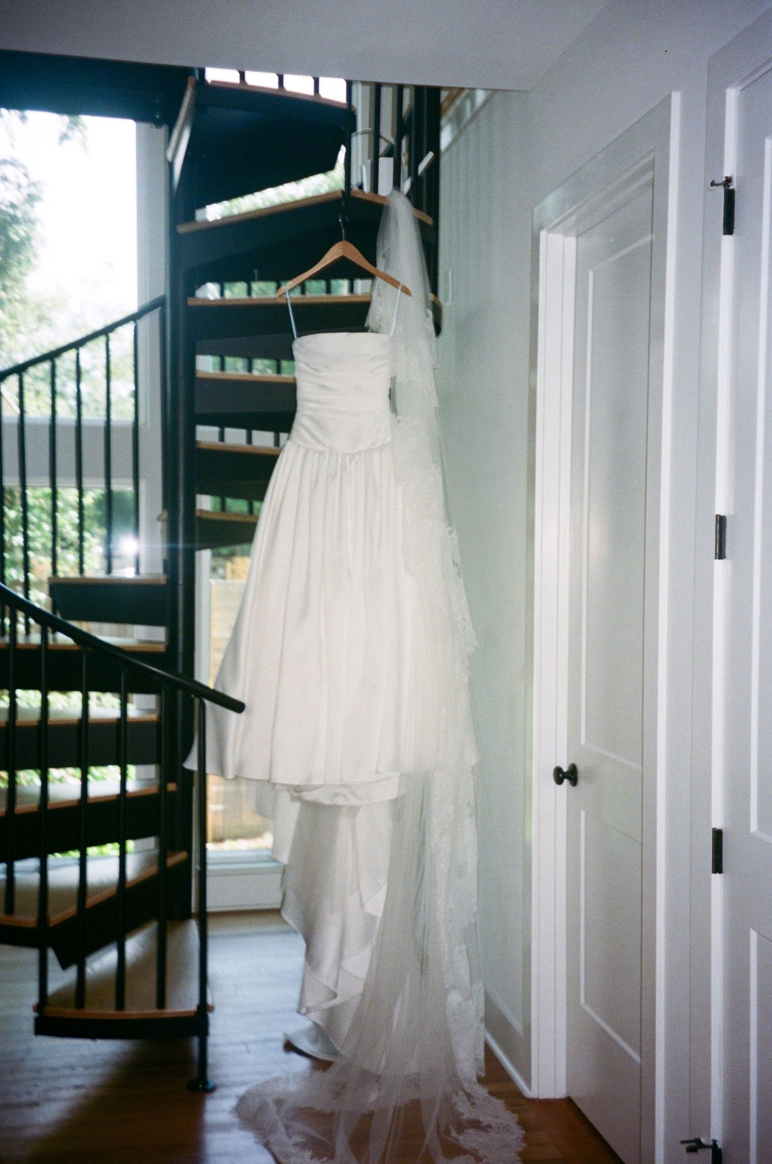 White wedding dress hanging in sunlit room with spiral staircase.