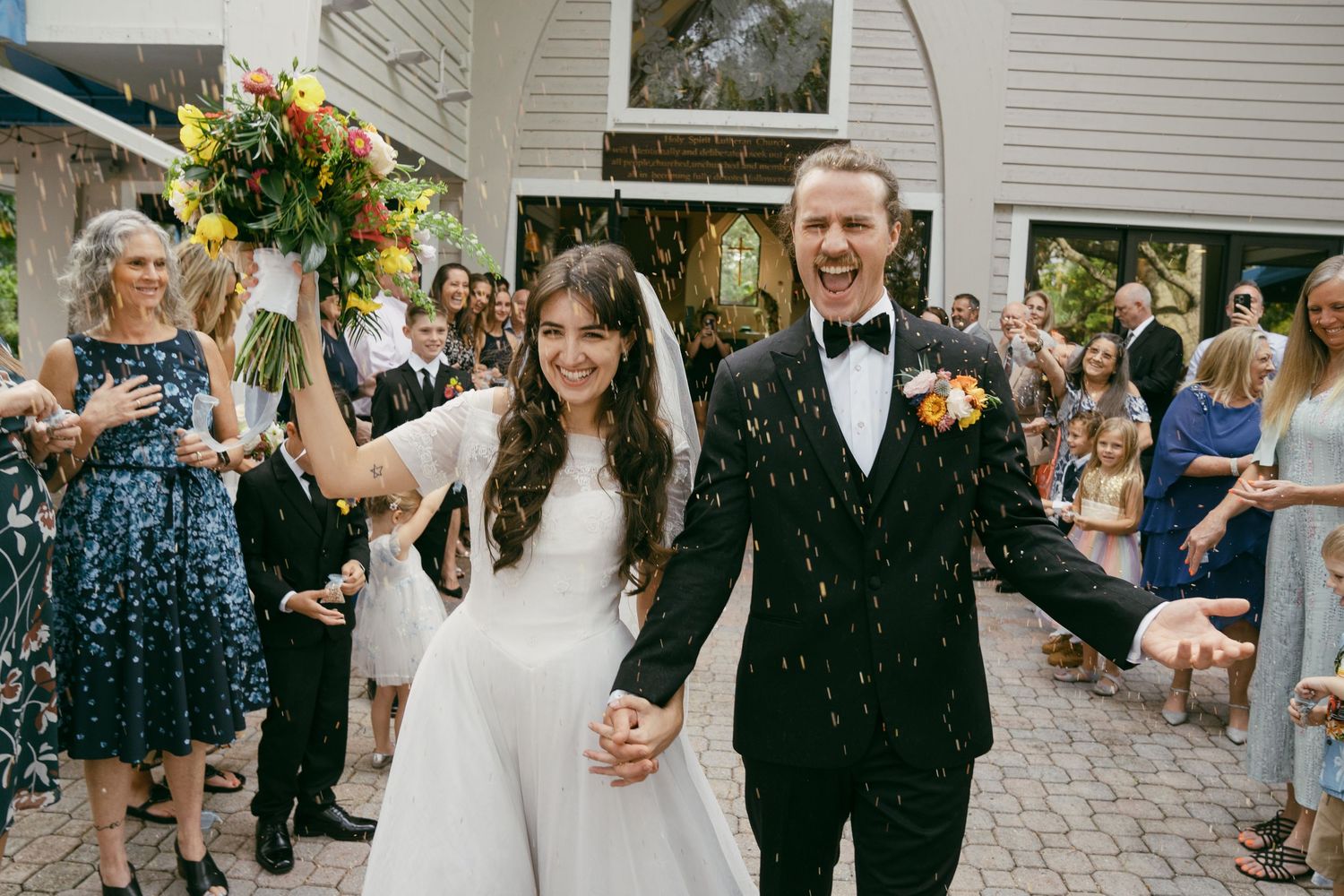 Wedding couple walks through a shower of confetti on a stone pathway surrounded by guests.