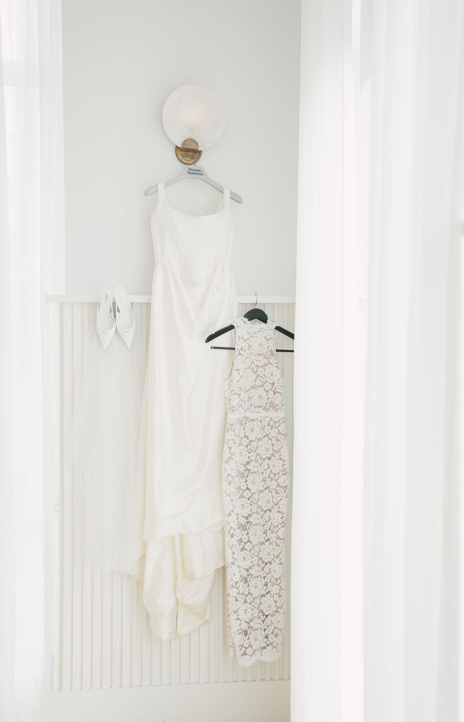 Four elegant white lace wedding dresses displayed hanging on hangers against a white curtained backdrop.
