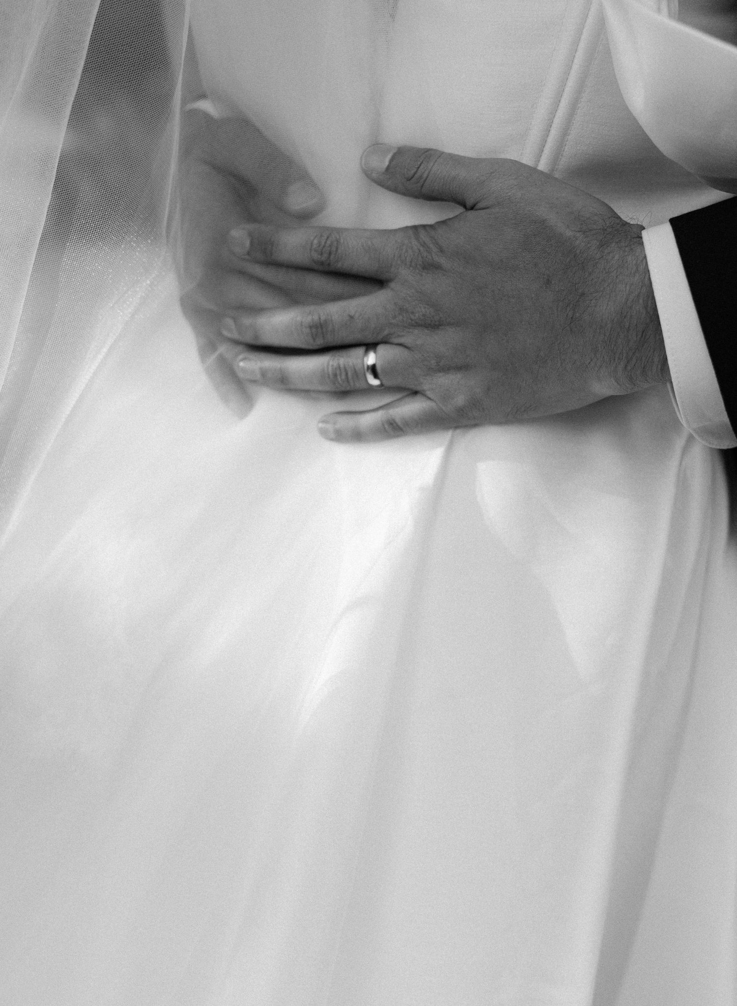Close-up of hands with wedding ring resting on white dress fabric in black and white.