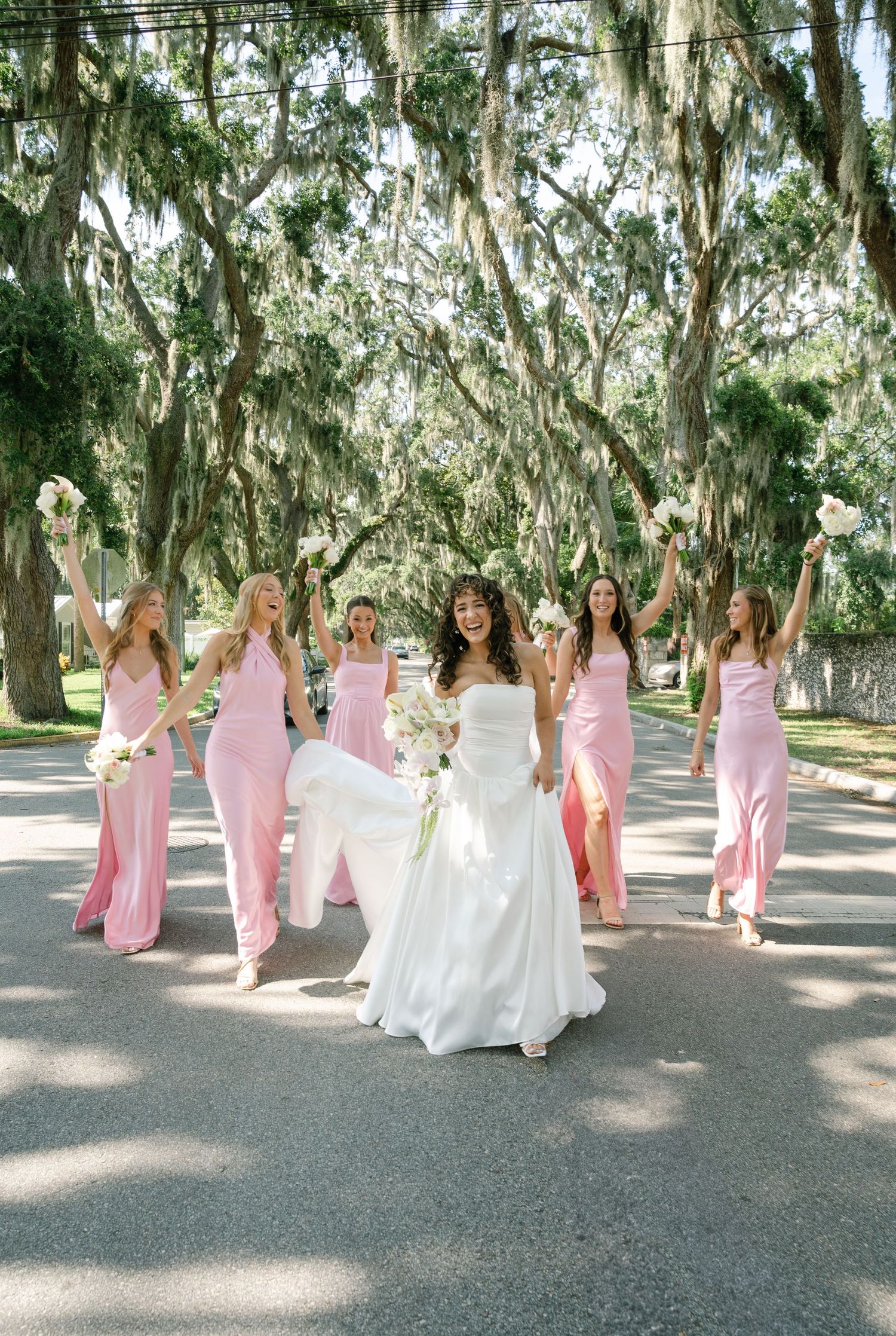 A bridal party poses on a tree-lined path with bridesmaids wearing pink dresses.