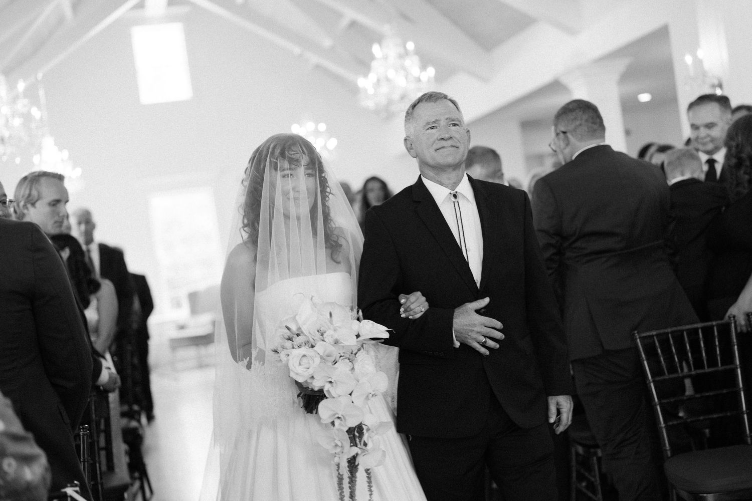 A bride and escort walk down the aisle in a bright white chapel with elegant chandeliers.