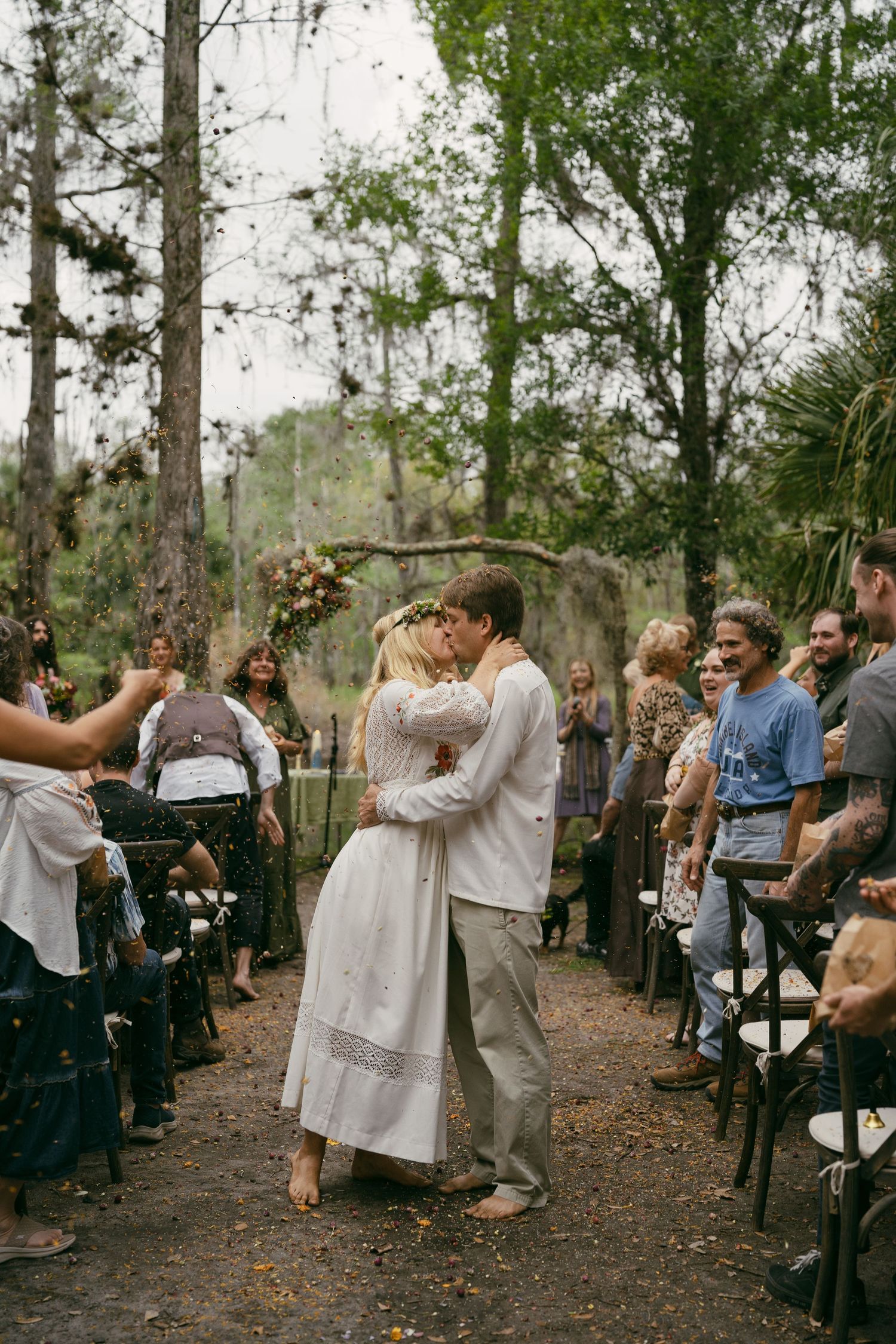 A romantic first kiss during a woodland wedding ceremony while guests watch from rustic wooden chairs.