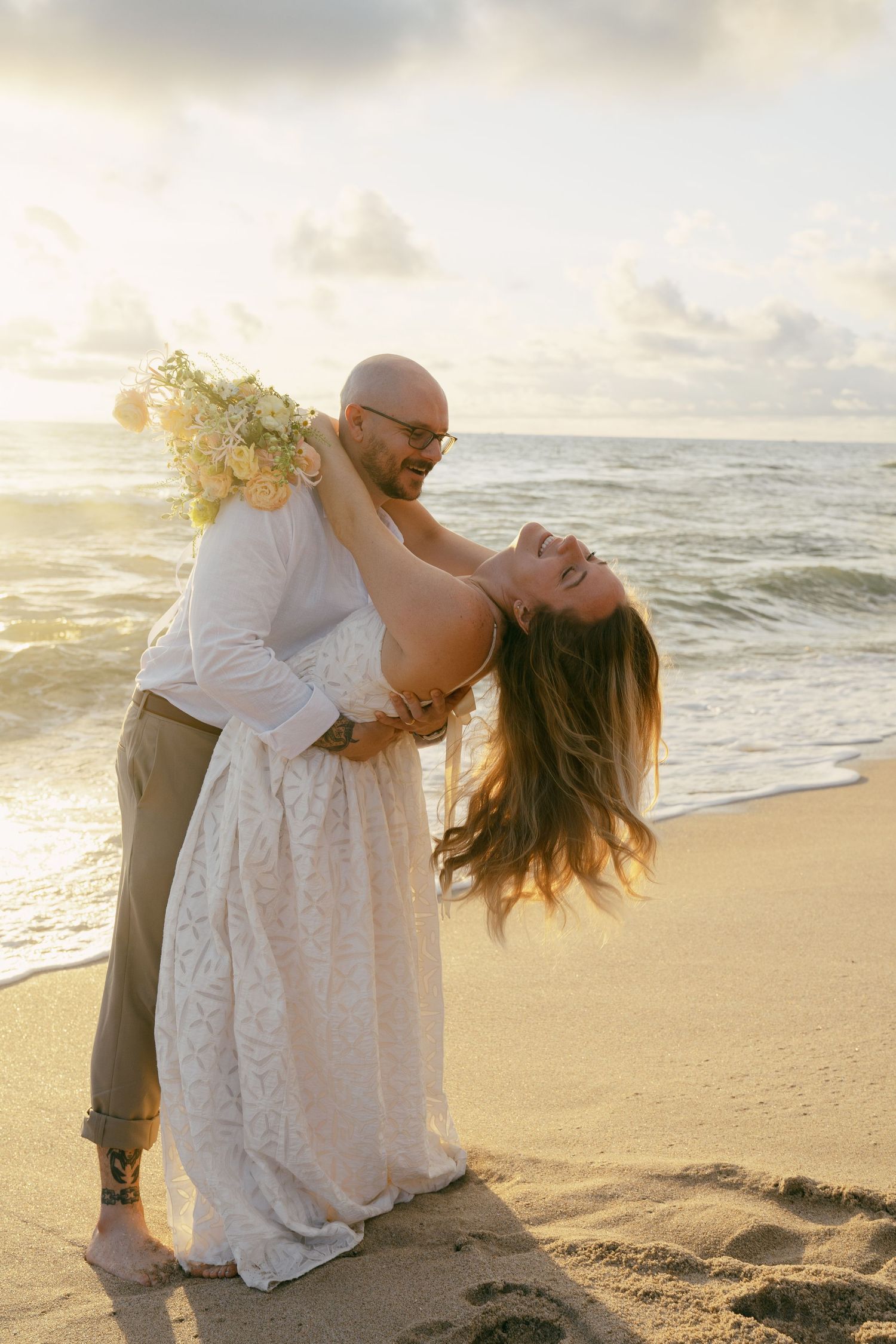 Romantic beach photoshoot with couple in white dress at sunset against ocean waves.