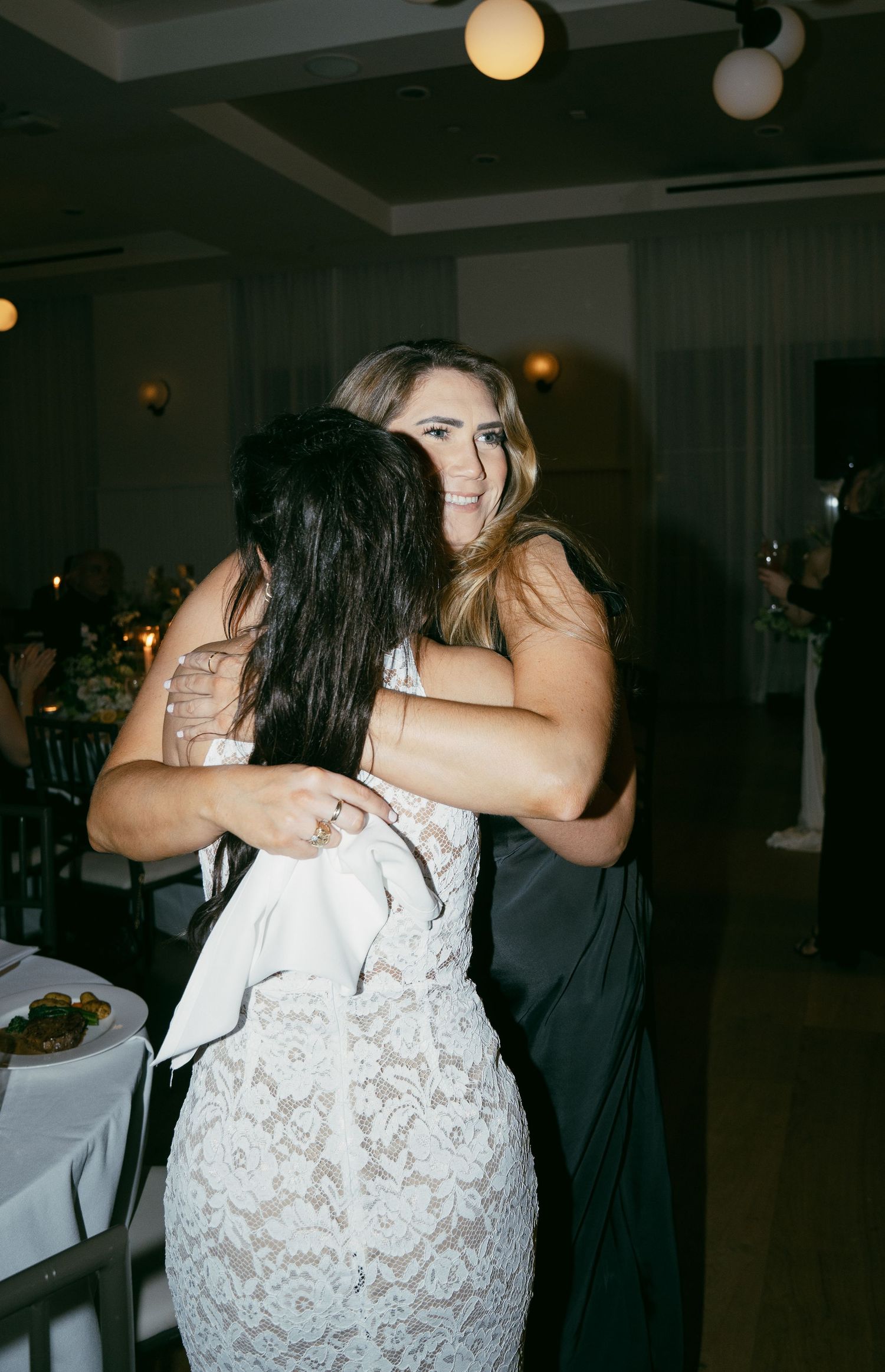 Two people share an emotional embrace at an indoor event with dimmed lighting and white tablecloths.
