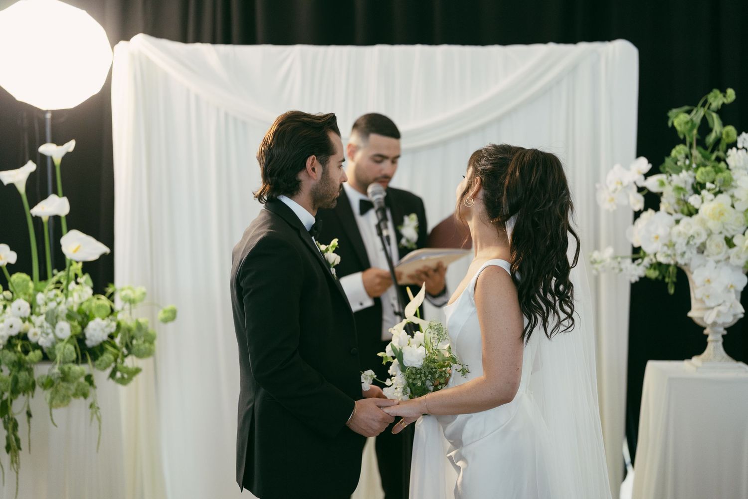 Wedding ceremony setup with white backdrop, floral arrangements, and couple exchanging vows.