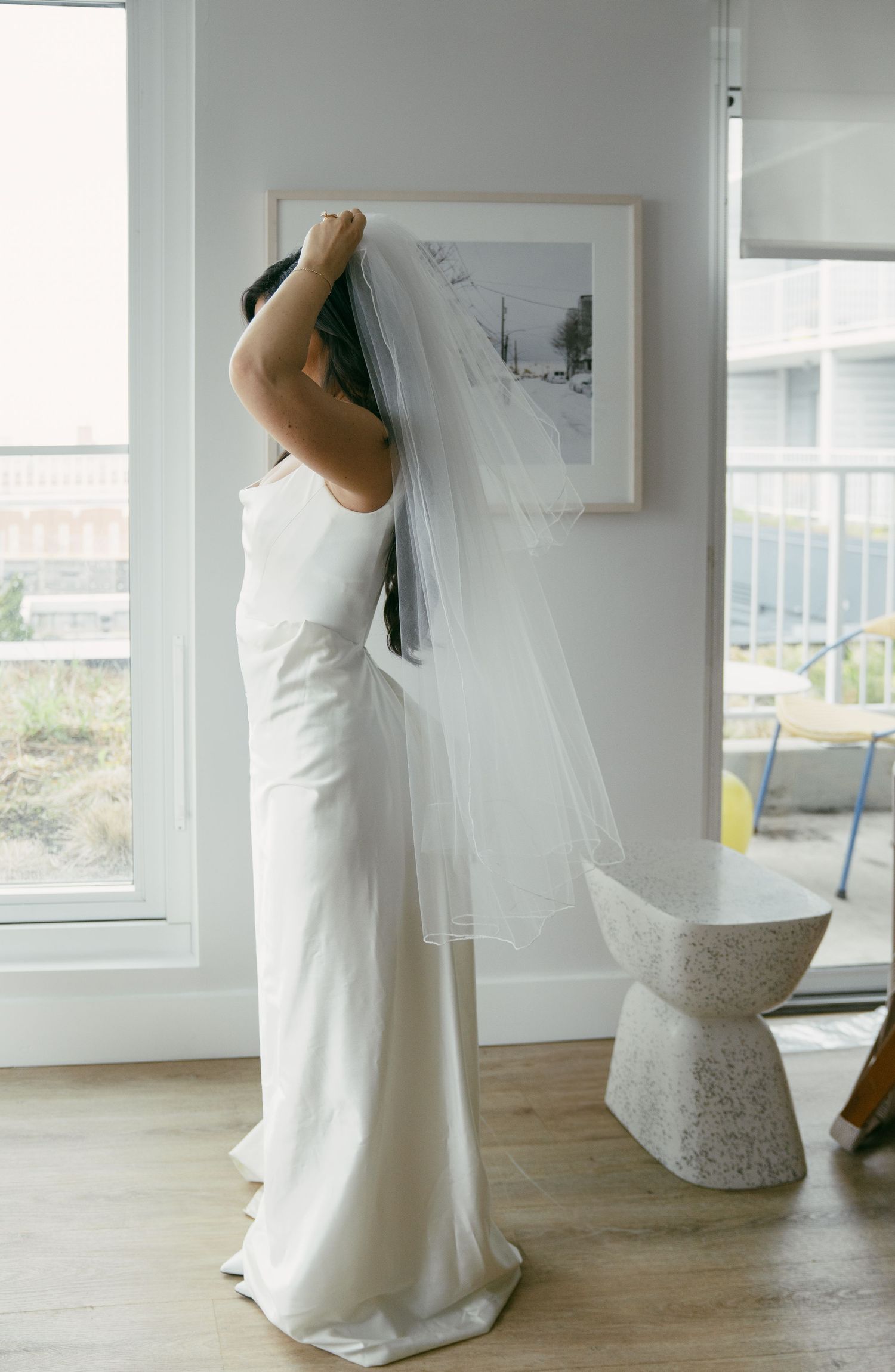 A bride in a sleek white wedding gown and flowing veil stands in a modern apartment with large windows.