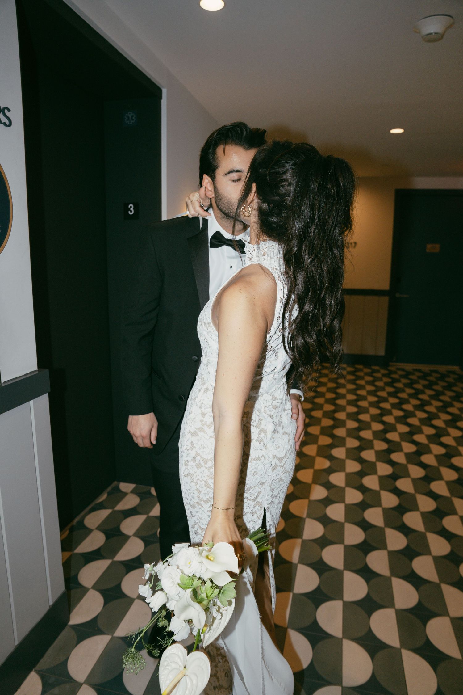 A couple kisses in a hotel hallway with a checkered floor pattern while the bride holds a white bouquet.