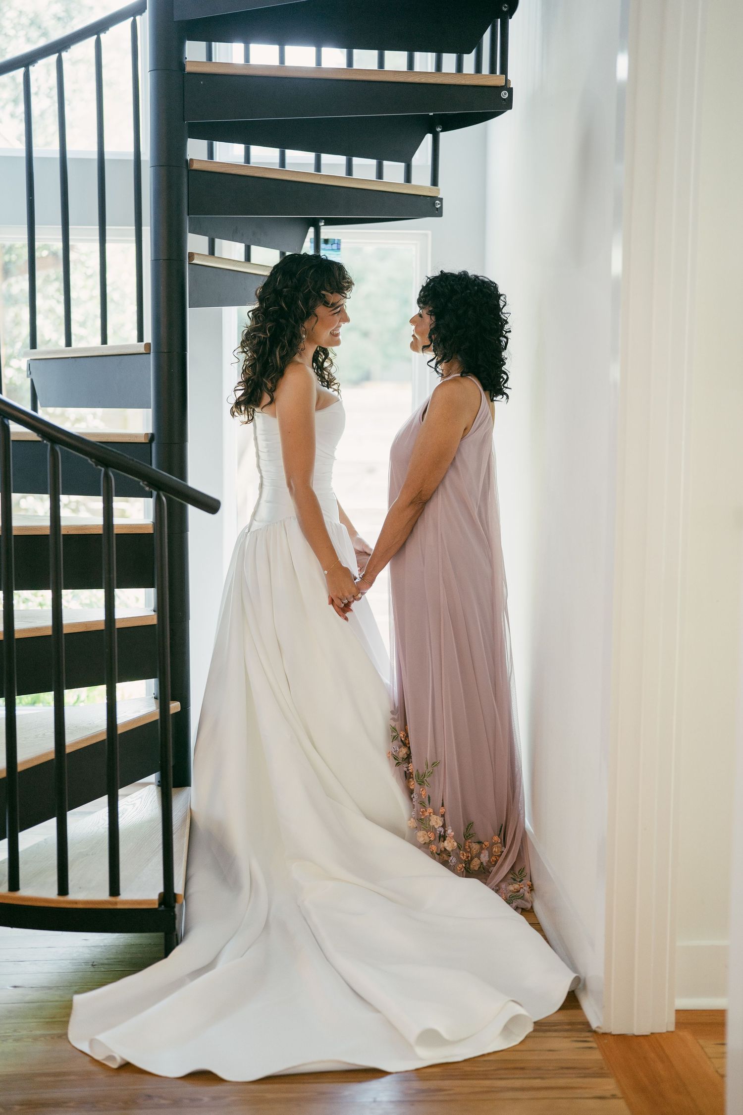 Two people in white and blush gowns standing together on a spiral staircase.