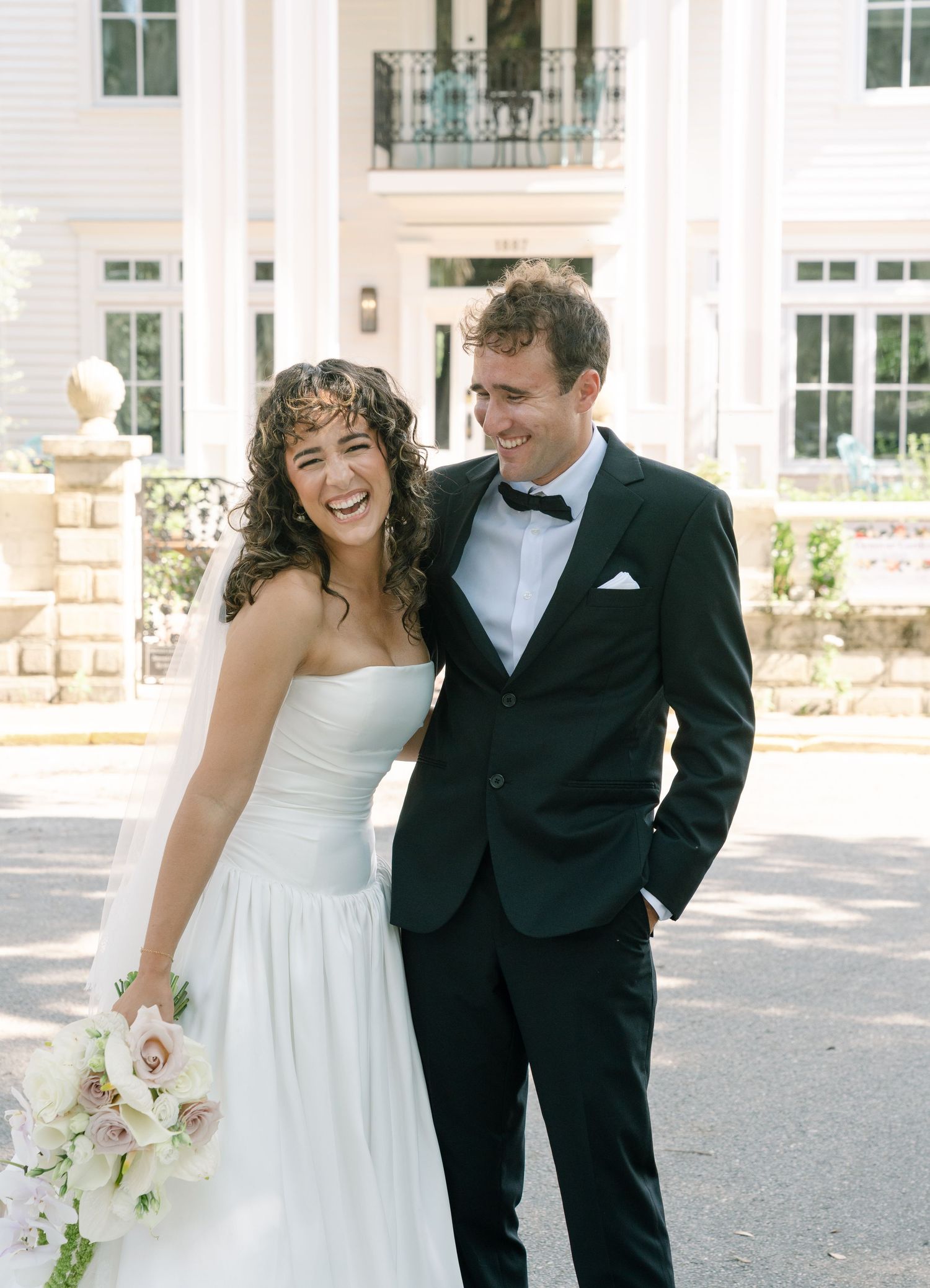 Newlyweds share a joyful moment outside an elegant white mansion.
