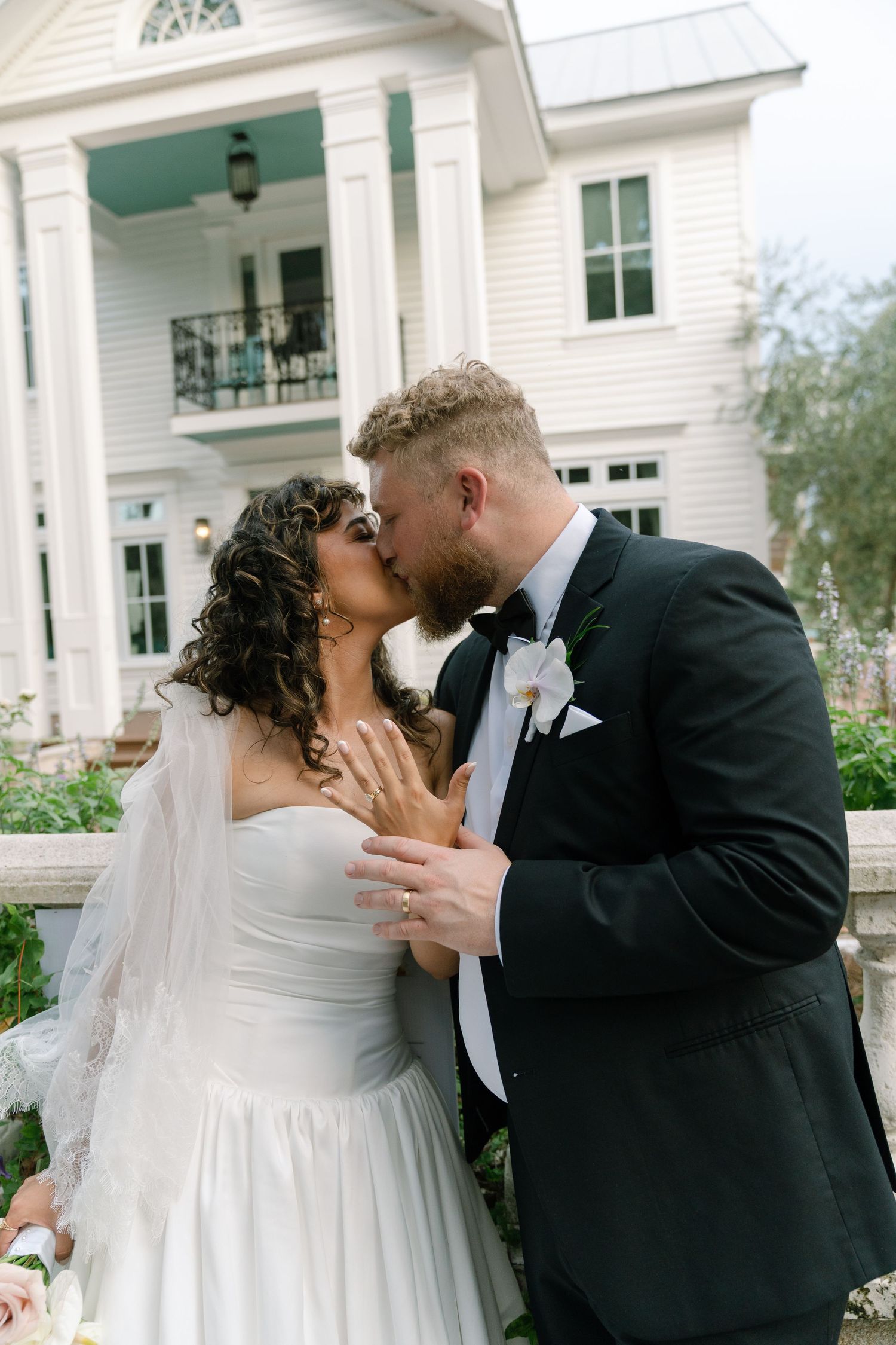 Newlyweds share an intimate kiss in front of a white Southern-style house.