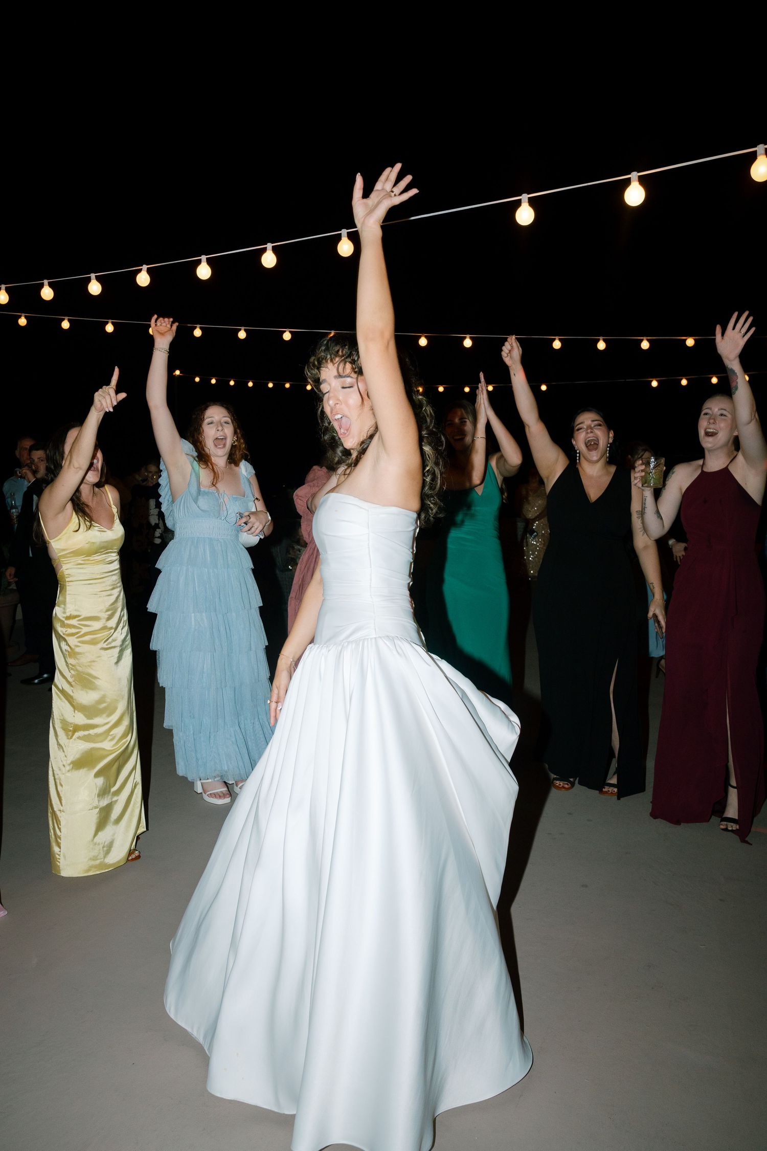 Wedding reception dance party under string lights with guests in colorful formal attire.