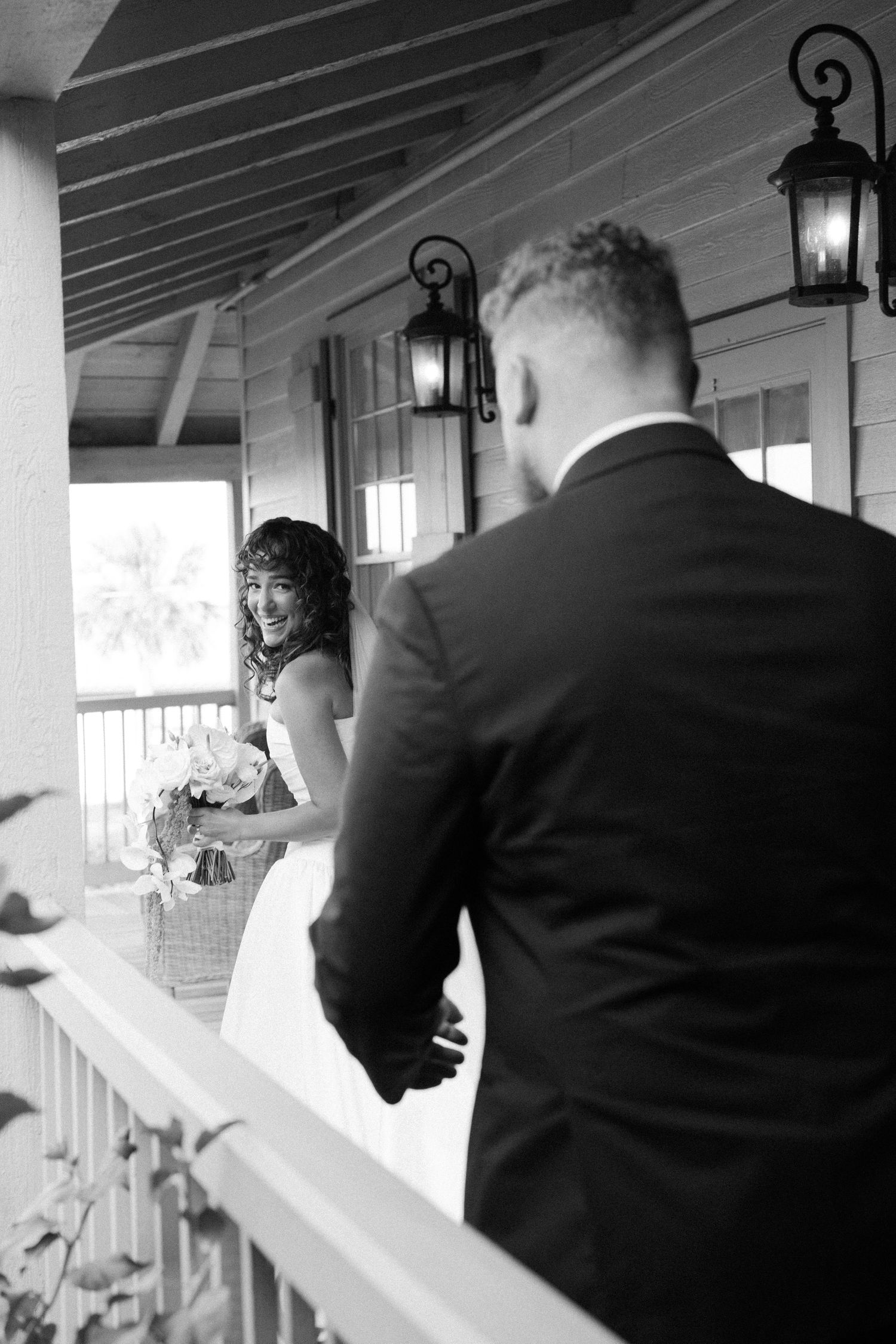 Black and white photo of newlyweds sharing a candid moment on a porch with vintage lanterns.