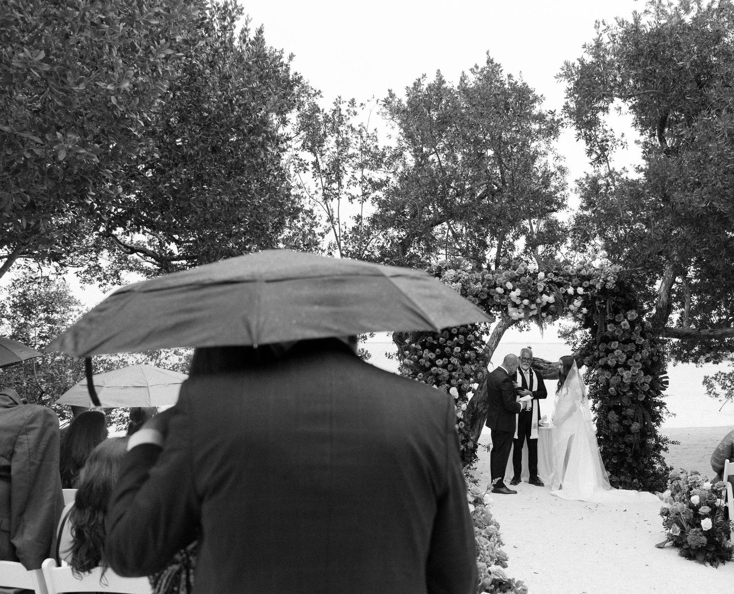 Guests with umbrellas watch an outdoor wedding ceremony under tree cover.