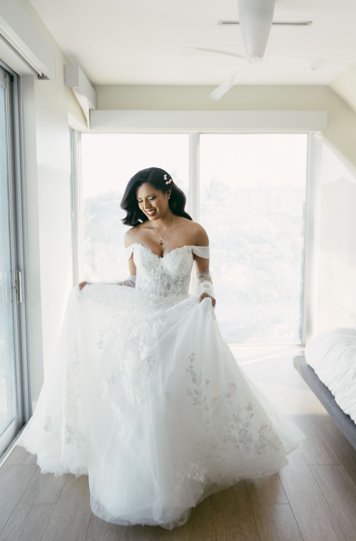 A bride twirls in an off-shoulder lace wedding gown near bright windows.