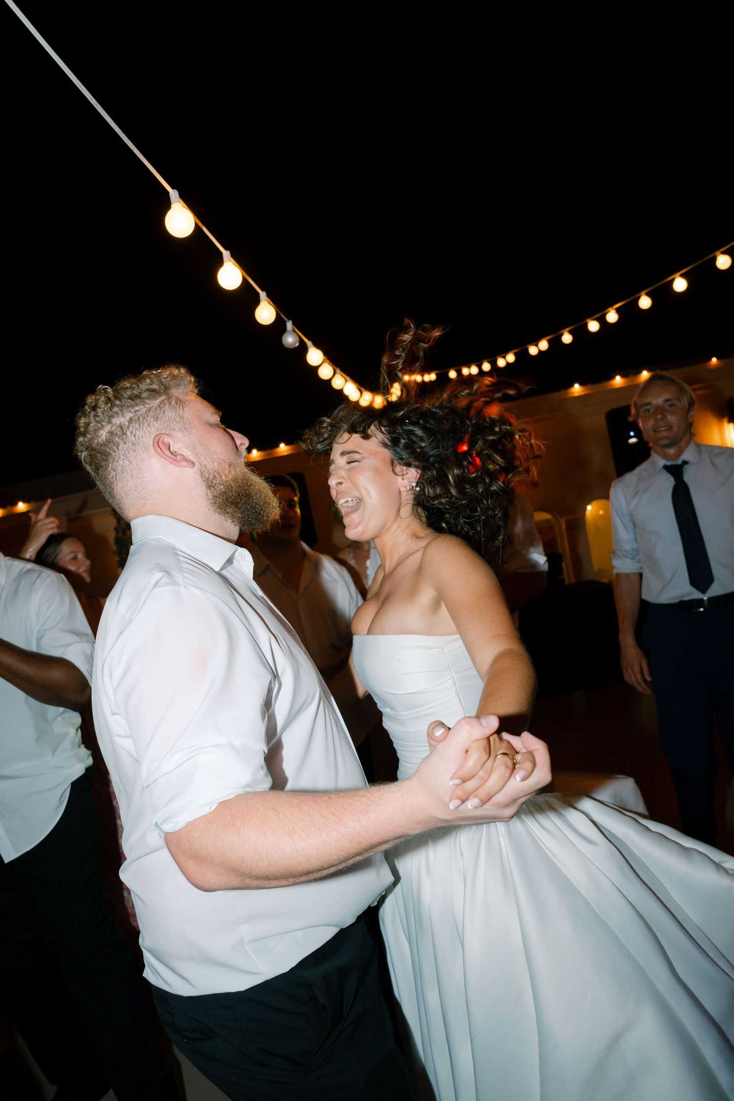 Newlyweds dancing and laughing under string lights at their evening reception.