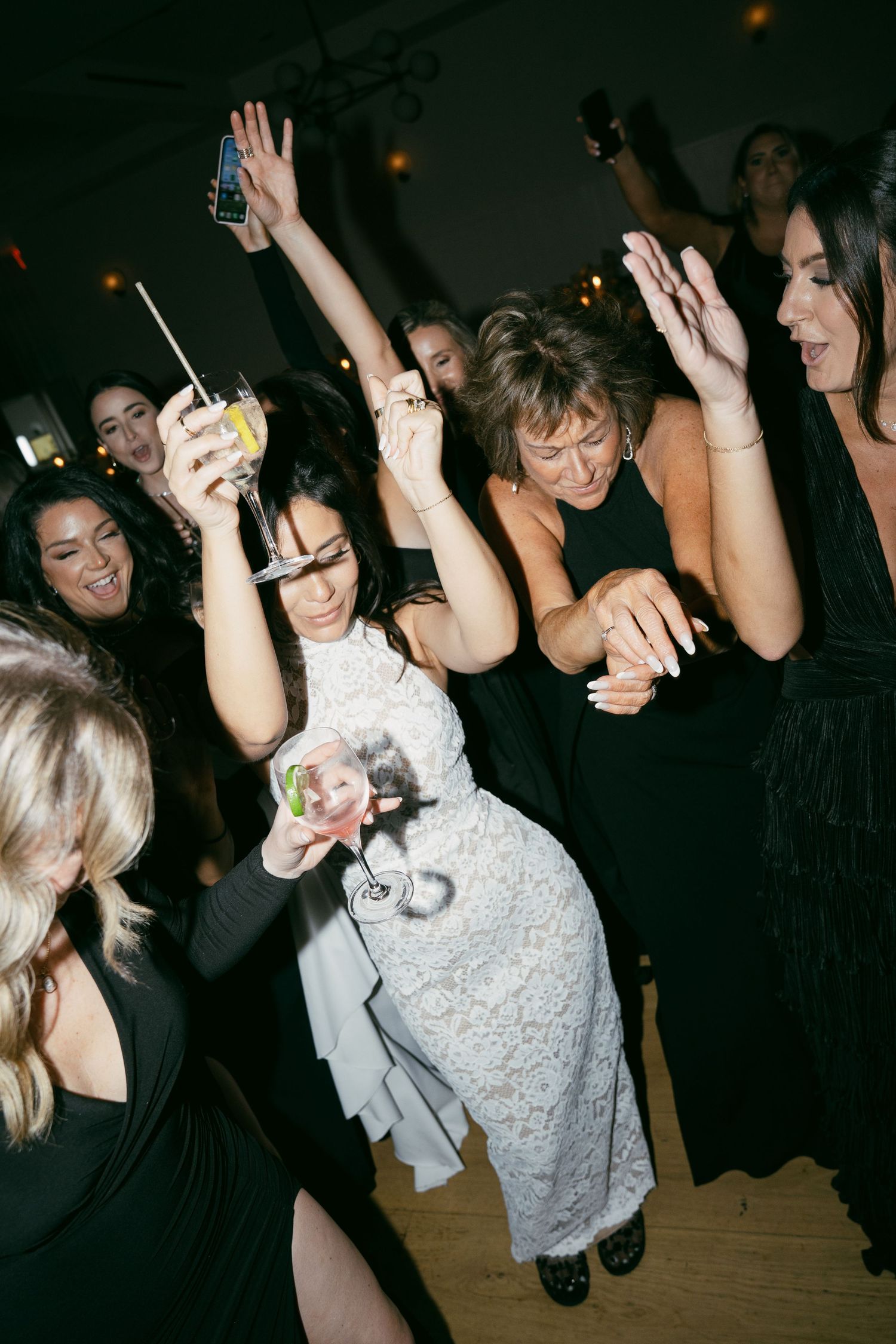 A group of party guests dance enthusiastically on a wooden dance floor, holding drinks and raising their hands in celebration.