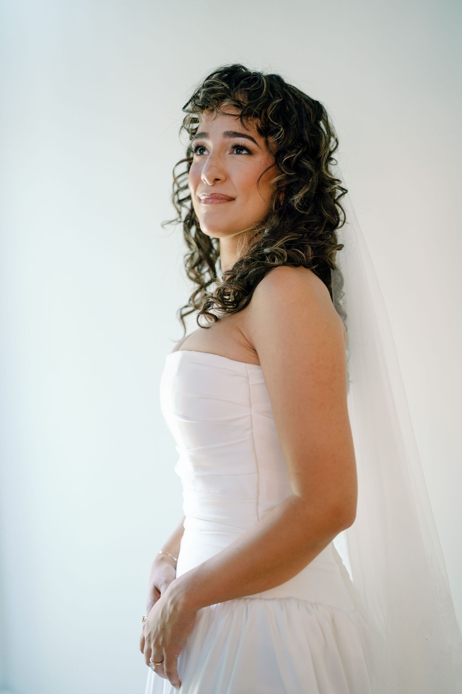 Bride in a strapless white wedding gown poses against a light background, showcasing her natural curly hair.