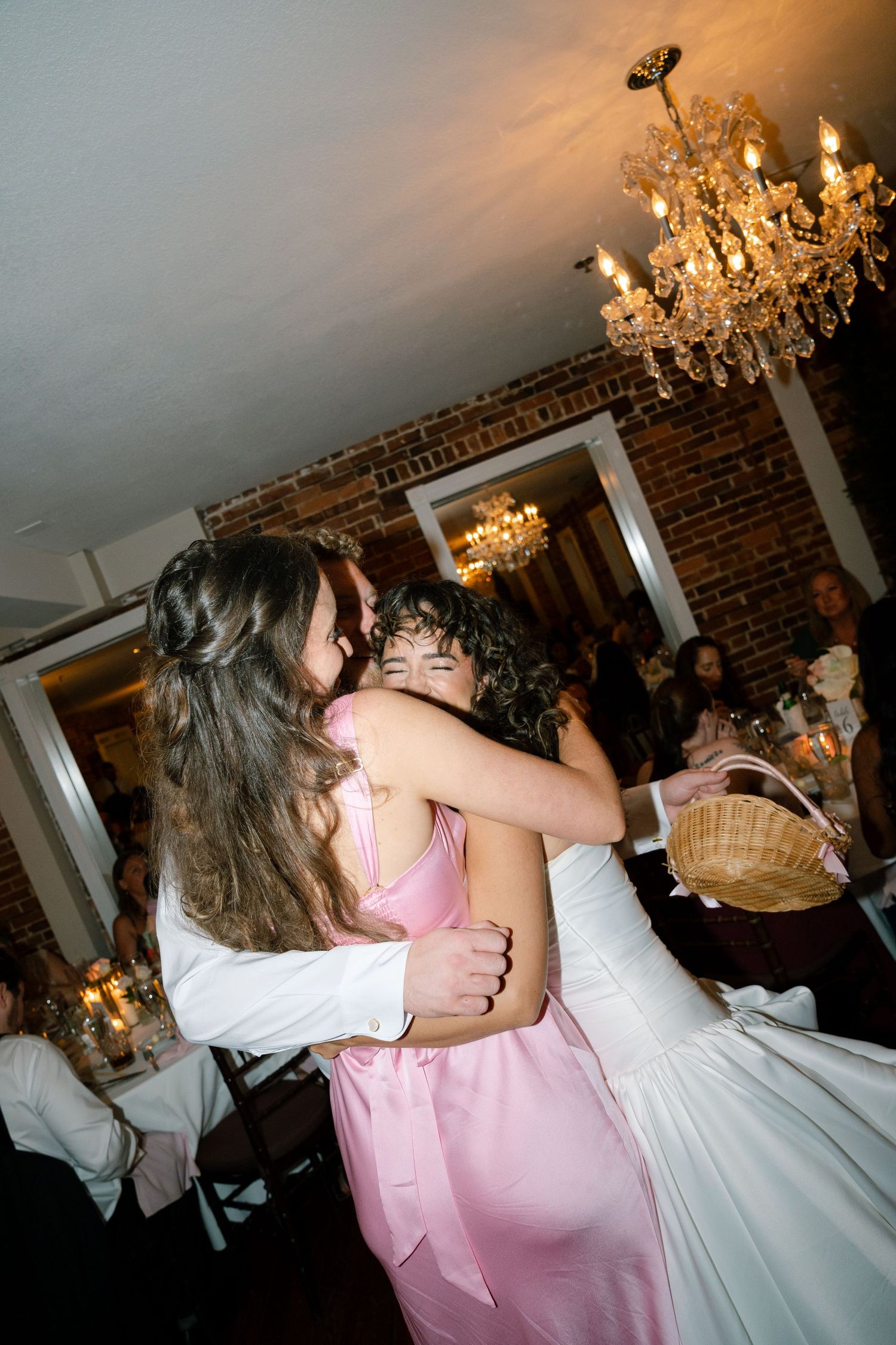 Pink-clad bridesmaids embracing the bride at a wedding reception with crystal chandeliers and brick walls.