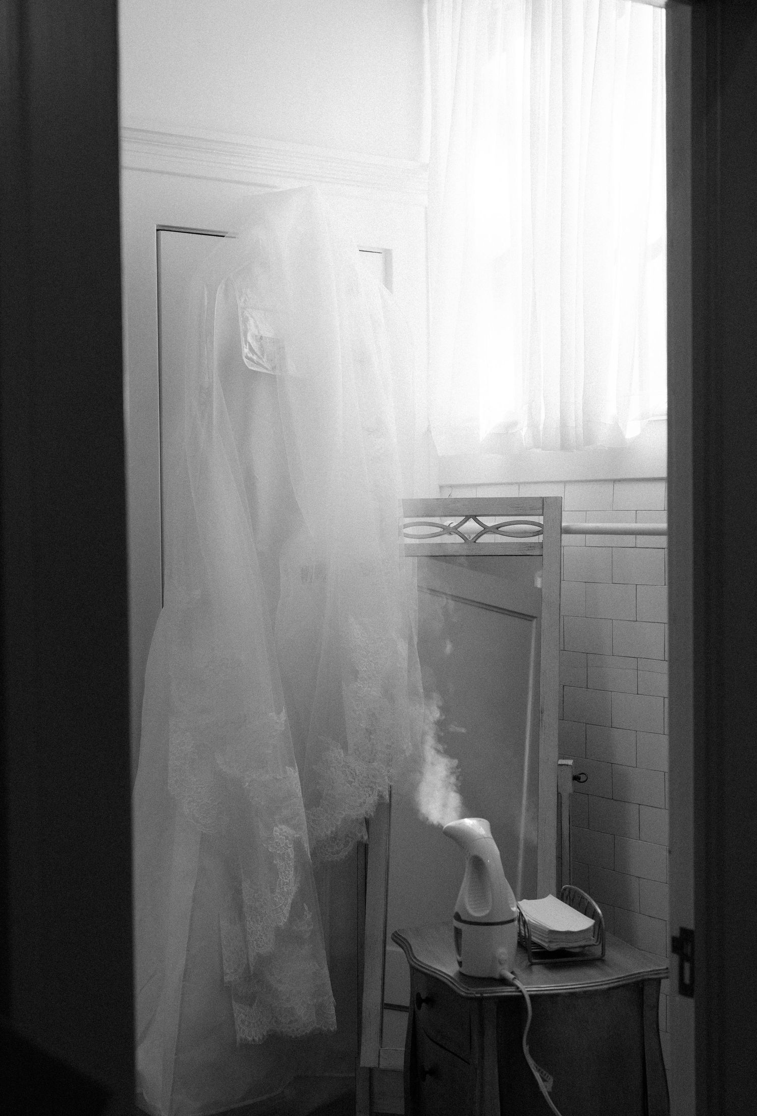 A white wedding dress hangs in a vintage bathroom with tiled walls and sheer curtains.