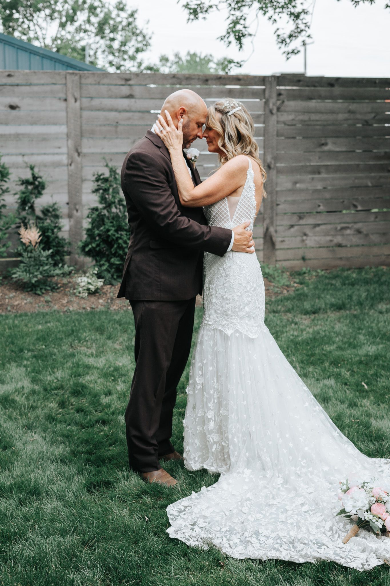 A couple in wedding attire share an intimate moment in a garden setting against a wooden fence.