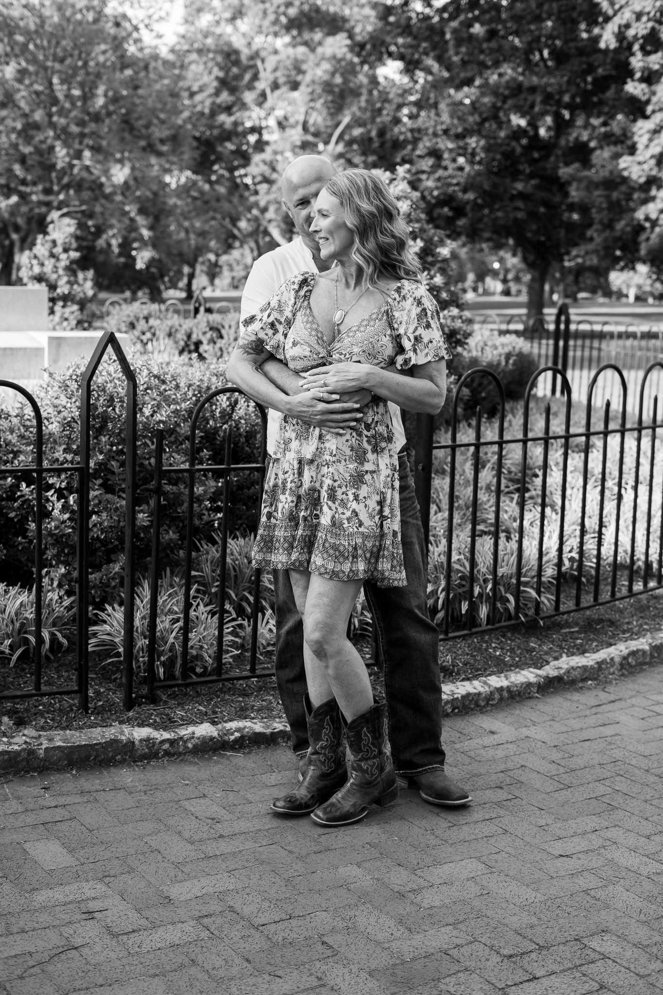 Black and white photograph of person in floral dress and boots standing against wrought iron fence in park setting.