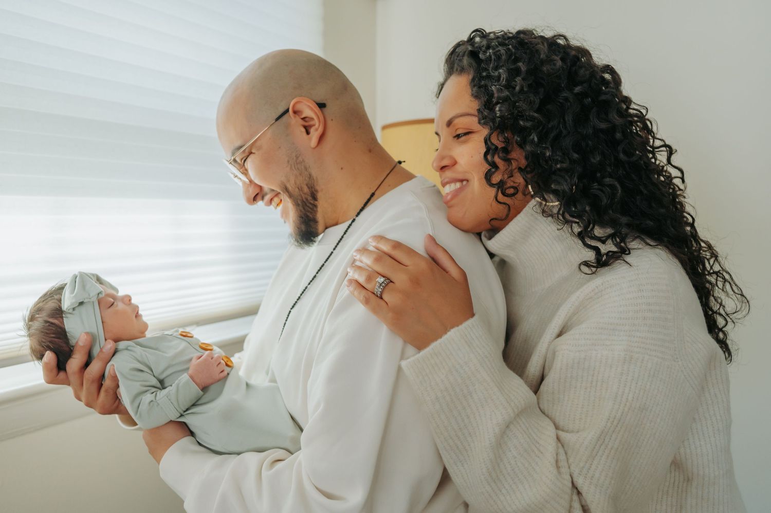 A couple in matching white robes shares a tender moment while holding a newborn baby in soft lighting.