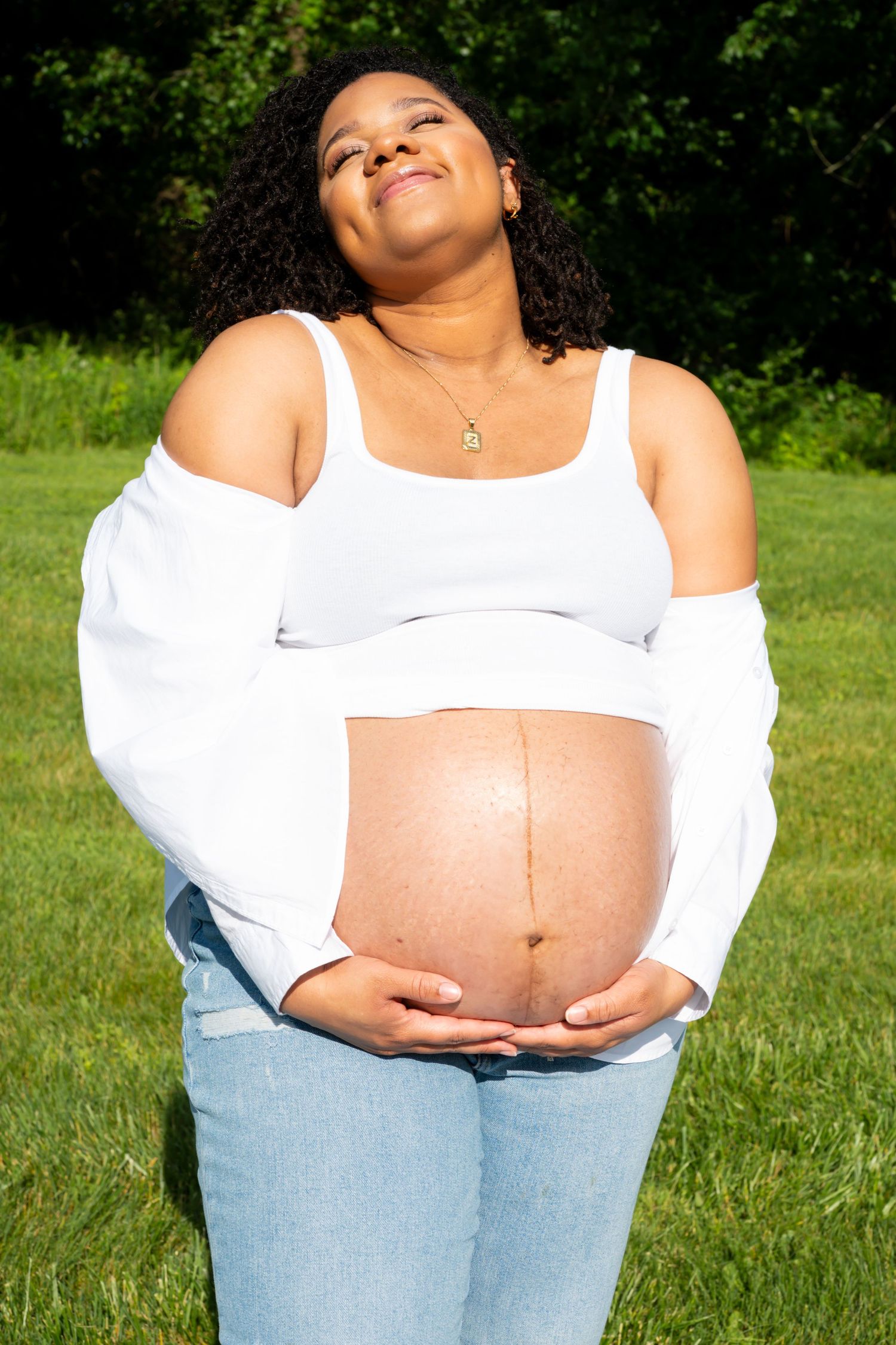 A pregnant person in a white crop top and light jeans poses outside on a sunny day in a grassy area.