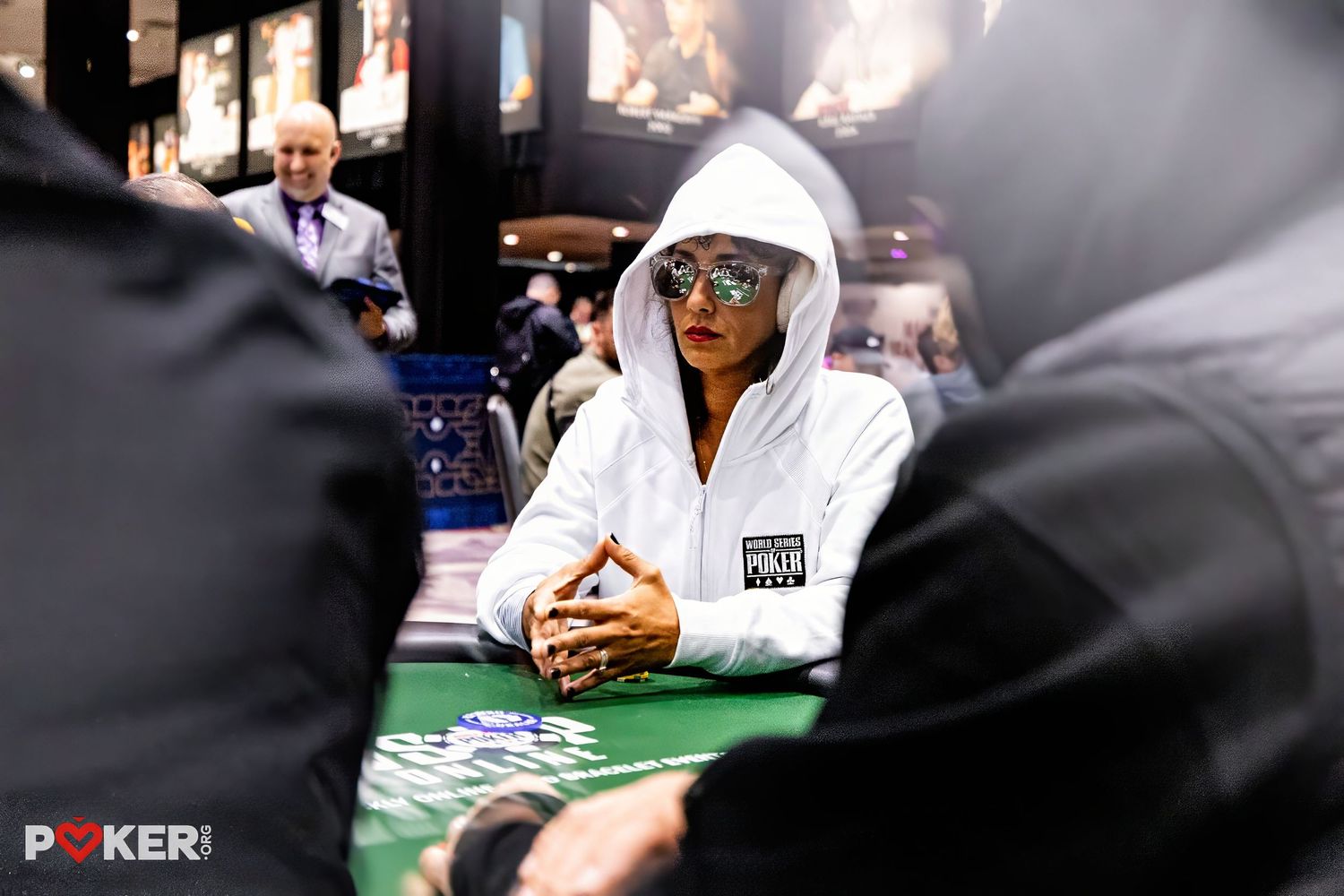 Player sits at poker table wearing white hoodie and glasses during poker tournament with dealer visible in background.