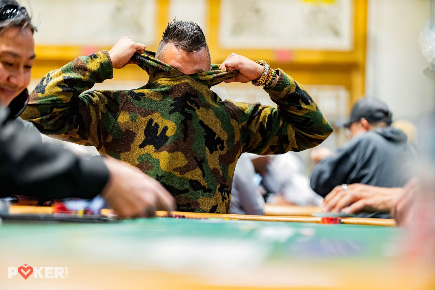 A person in camouflage military attire holds their head while sitting at a card gaming table.