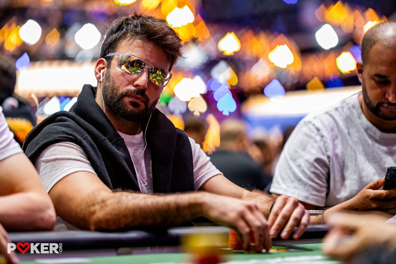 Players seated at poker tournament tables with colorful bokeh lighting in background.