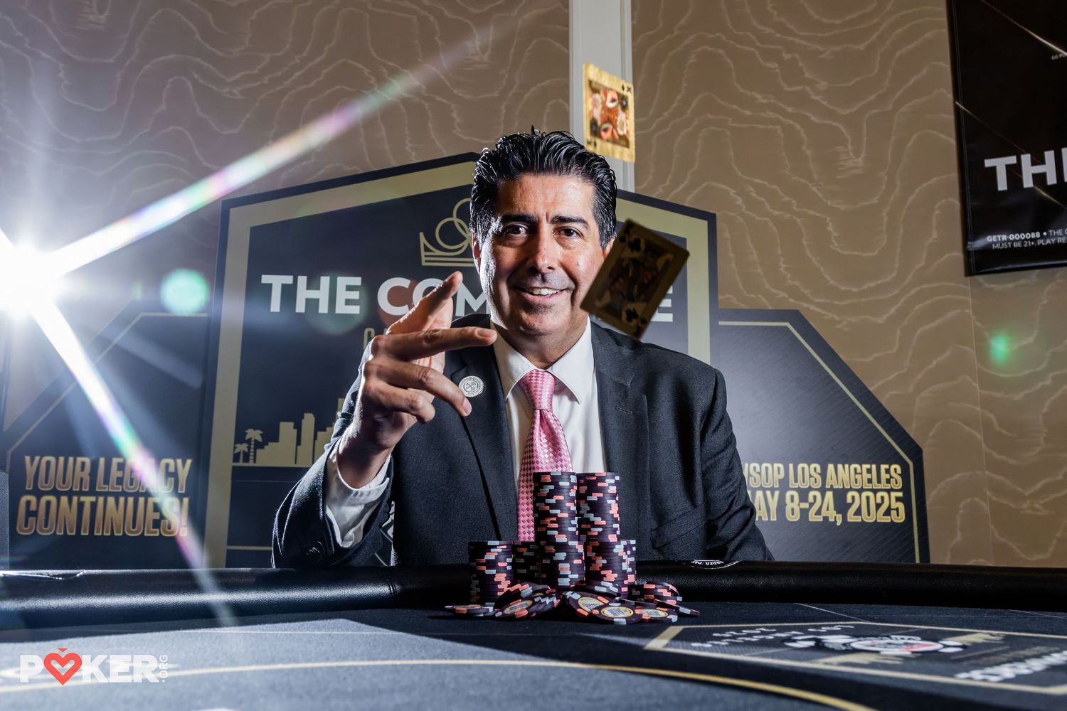 Professional poker player sits at casino table with stack of chips while holding glass of whiskey in elegant suit.