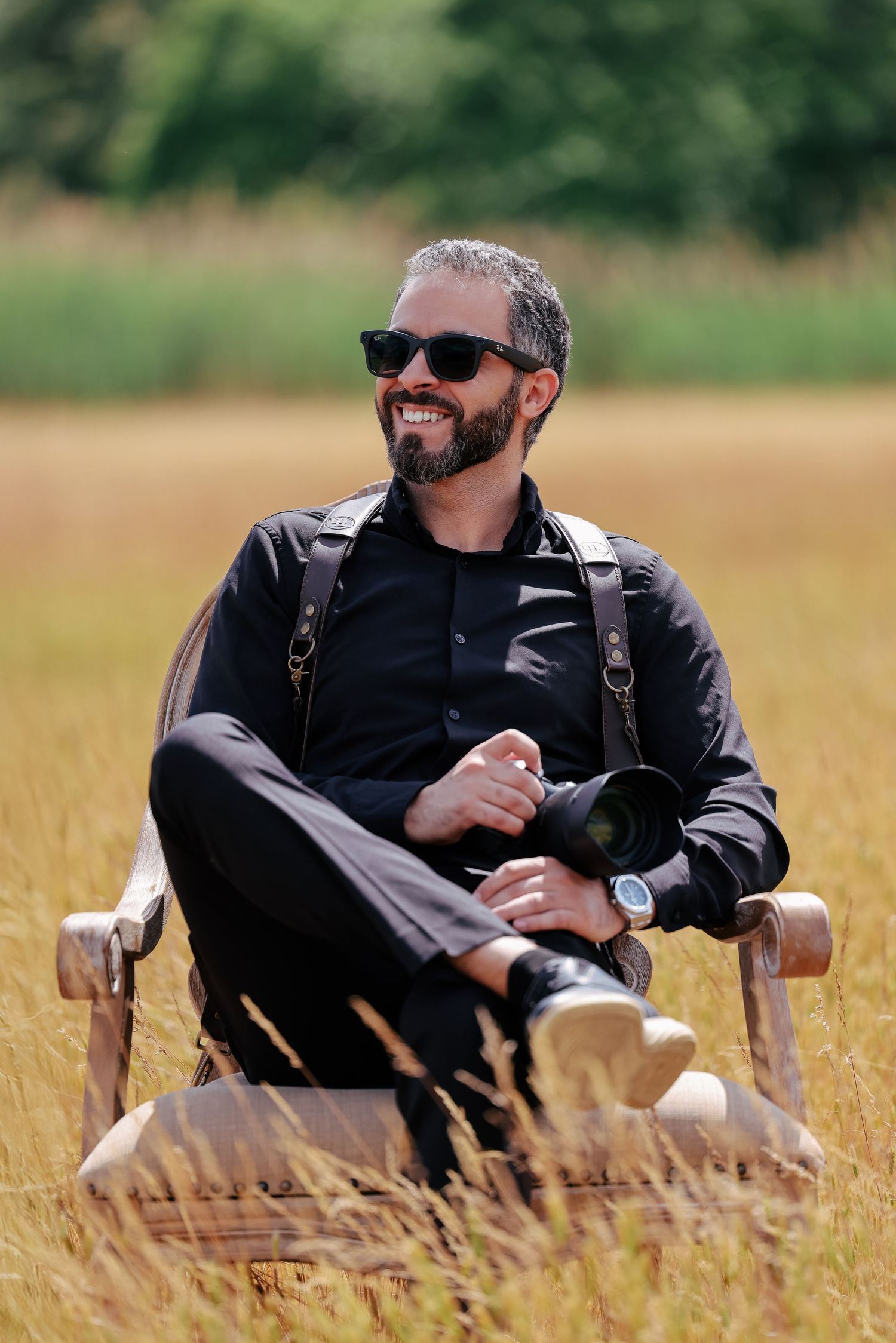 Man in black attire and sunglasses relaxes in wooden chair in sunny wheat field.