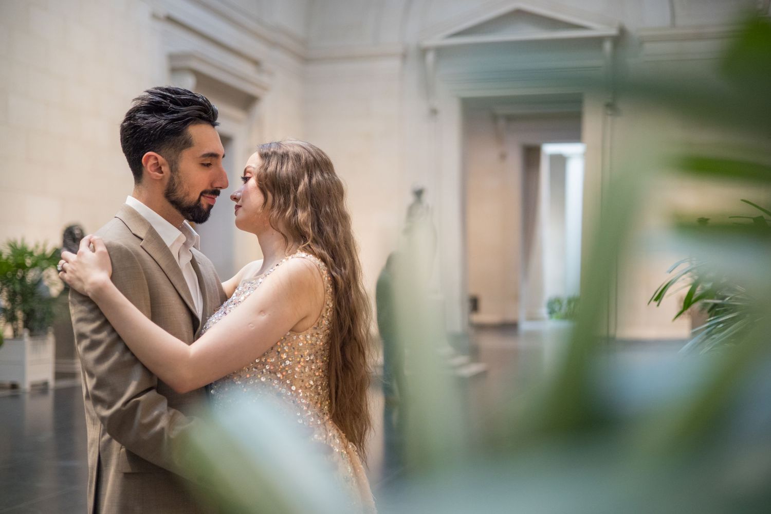 A couple in formal wear shares an intimate embrace against a light colored wall.